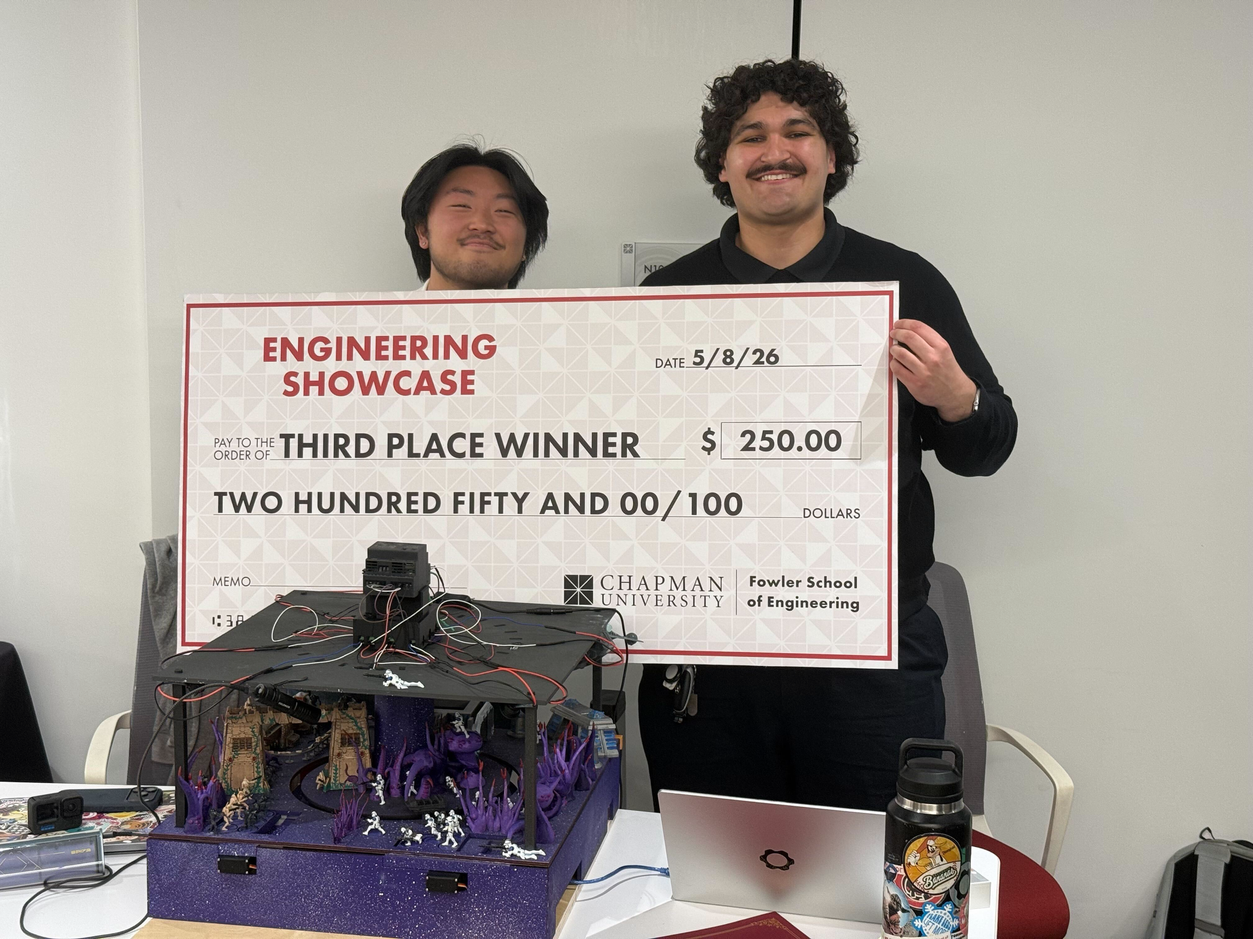 Two men standing behind a table with a large sign that says 'Engineering Showcase' and an award amount of $250.00. The table has a purple display with small robotic figures and electronic components. The man on the right has curly hair and is smiling, while the man on the left has straight hair and is smiling with his eyes closed.