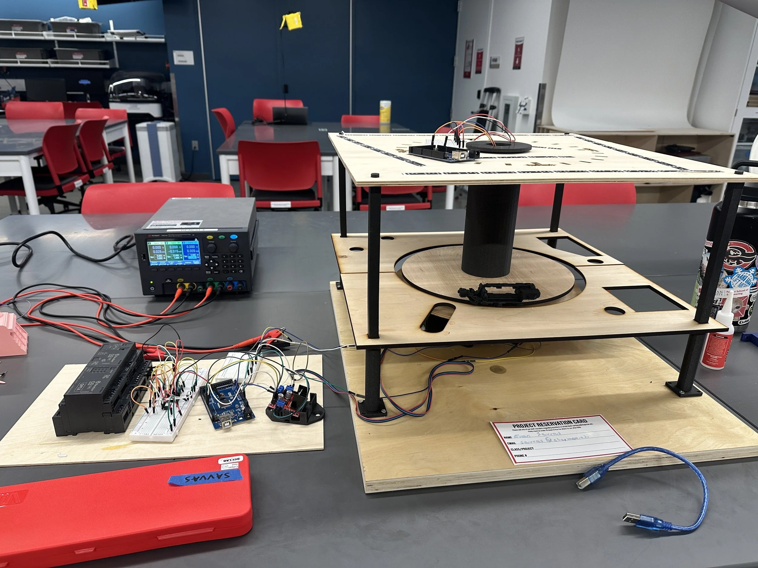 Workstation with electronic components and wires, including a breadboard, microcontroller, and power supply, set up on tables and a wooden platform in a classroom or lab environment.