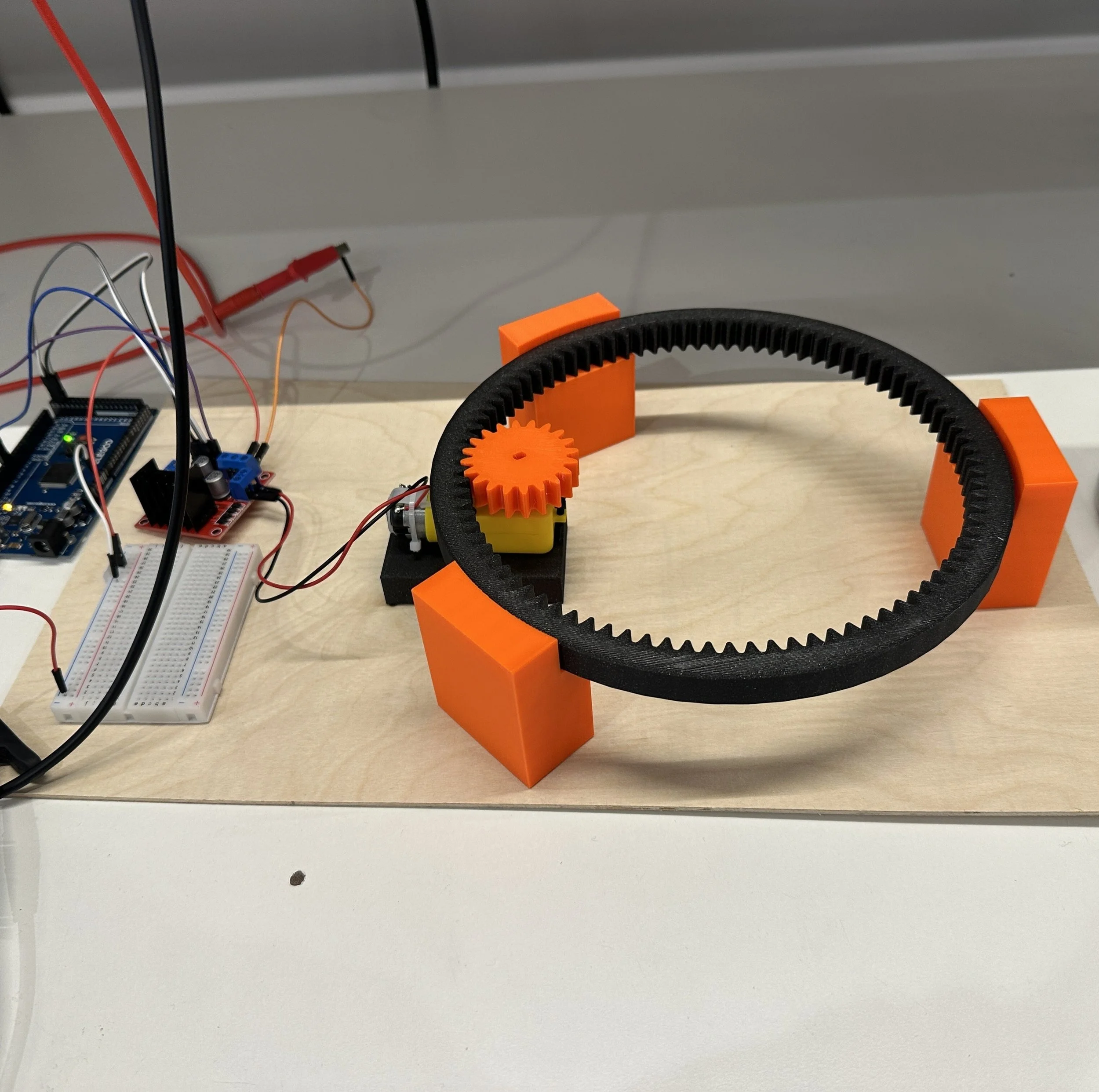 A small robotic setup on a white table with electronic components and wires, including a breadboard, Arduino, and a belt-drive mechanism with orange and black gears.