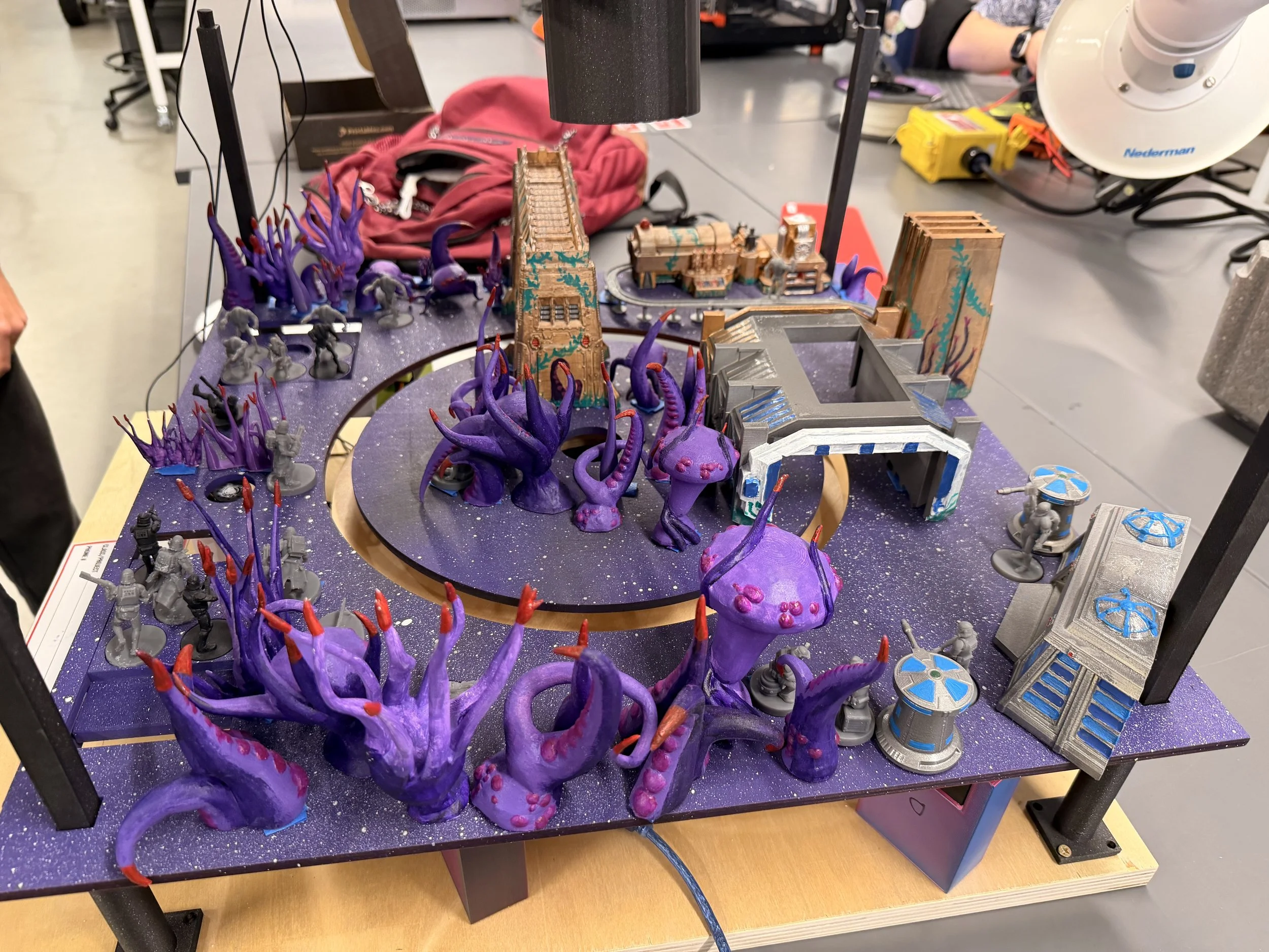 Tabletop gaming terrain with purple tentacle-like structures, miniature buildings, and scattered miniature figurines on a speckled purple surface.