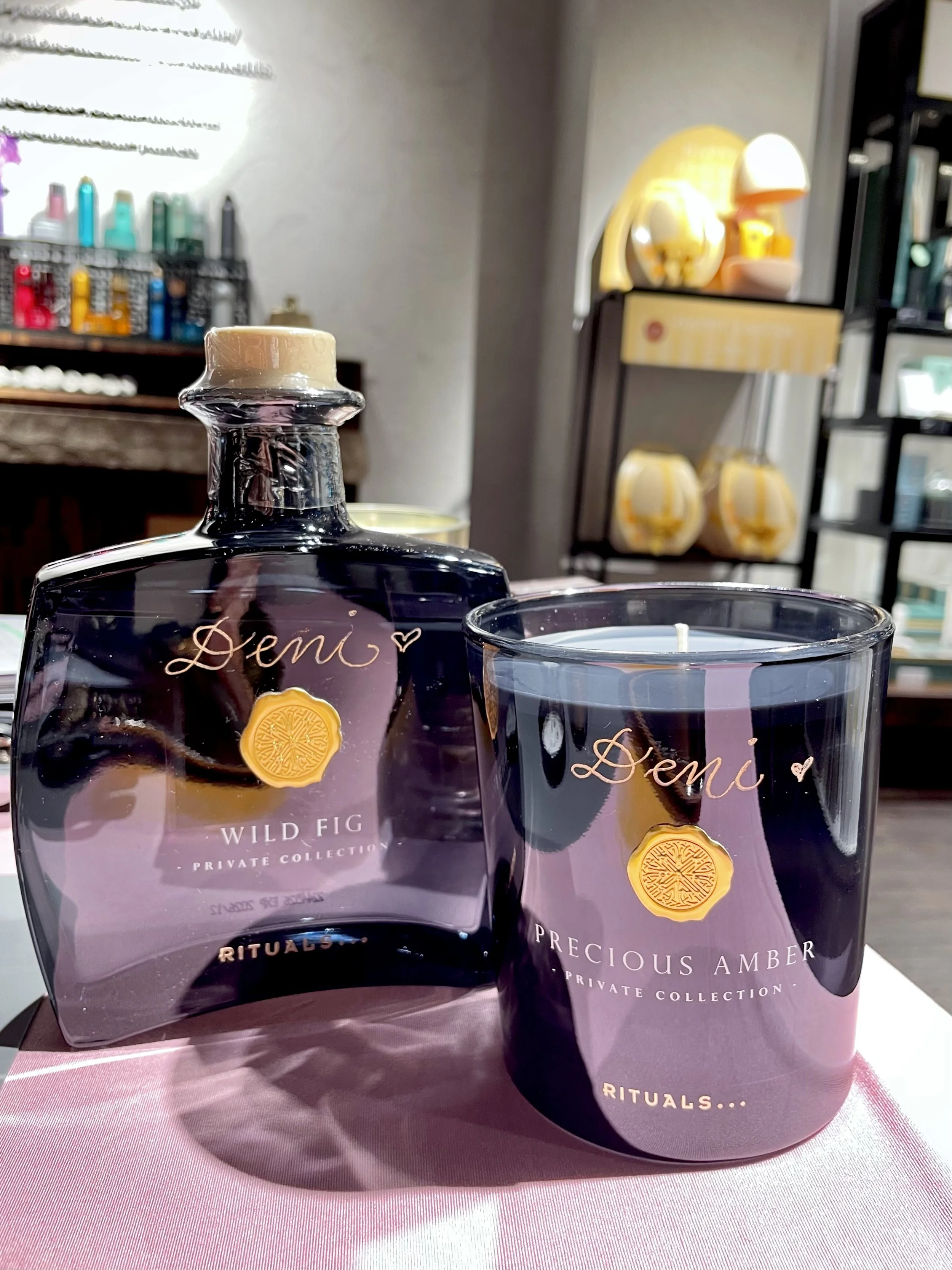 A bottle of "Wild Fig" fragrance and a "Precious Amber" candle from the Private Collection by Rituals displayed on a table.