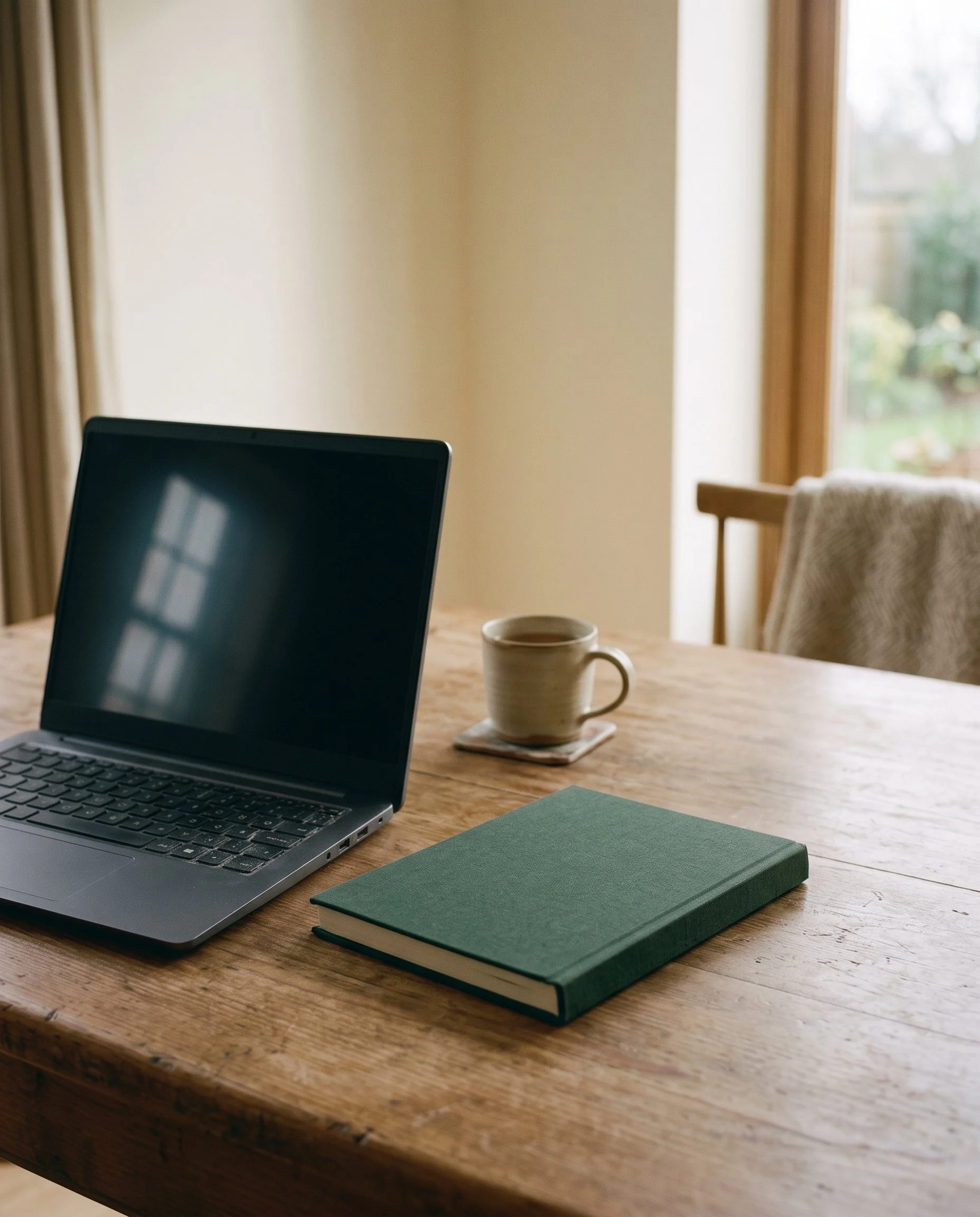 An open laptop, closed green notebook and mug of tea on a wooden table in morning light