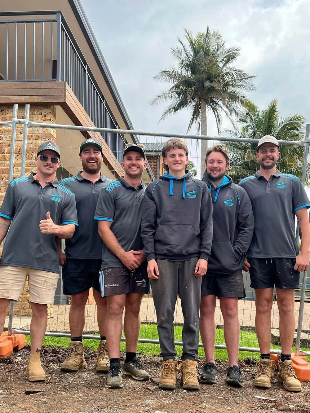 Group of six men wearing work uniforms and boots standing in front of a fenced construction site with a building and palm trees in the background.