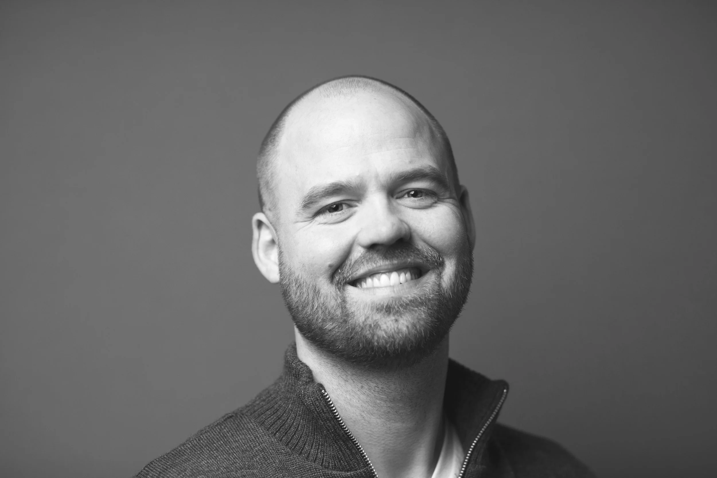 Sort-hvid portrætfotos af en mand med skæg og smil, iført en mørk sweater mod en simpel baggrund.