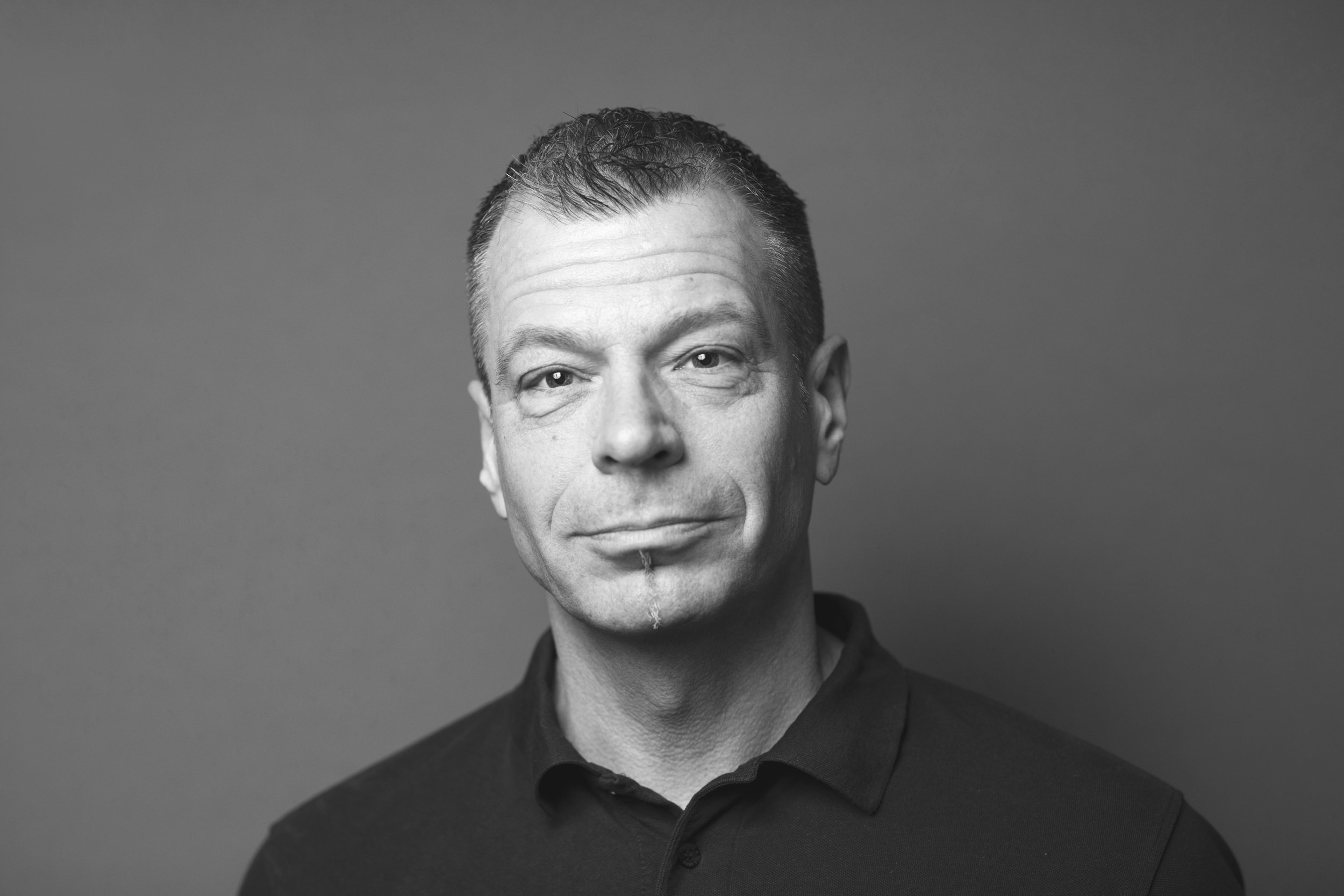 Sort-hvid portrætfoto af en mand med kort, mørkt hår, let skægstub og en selvsikker udstråling, iført sort polo i en studieindstilling.