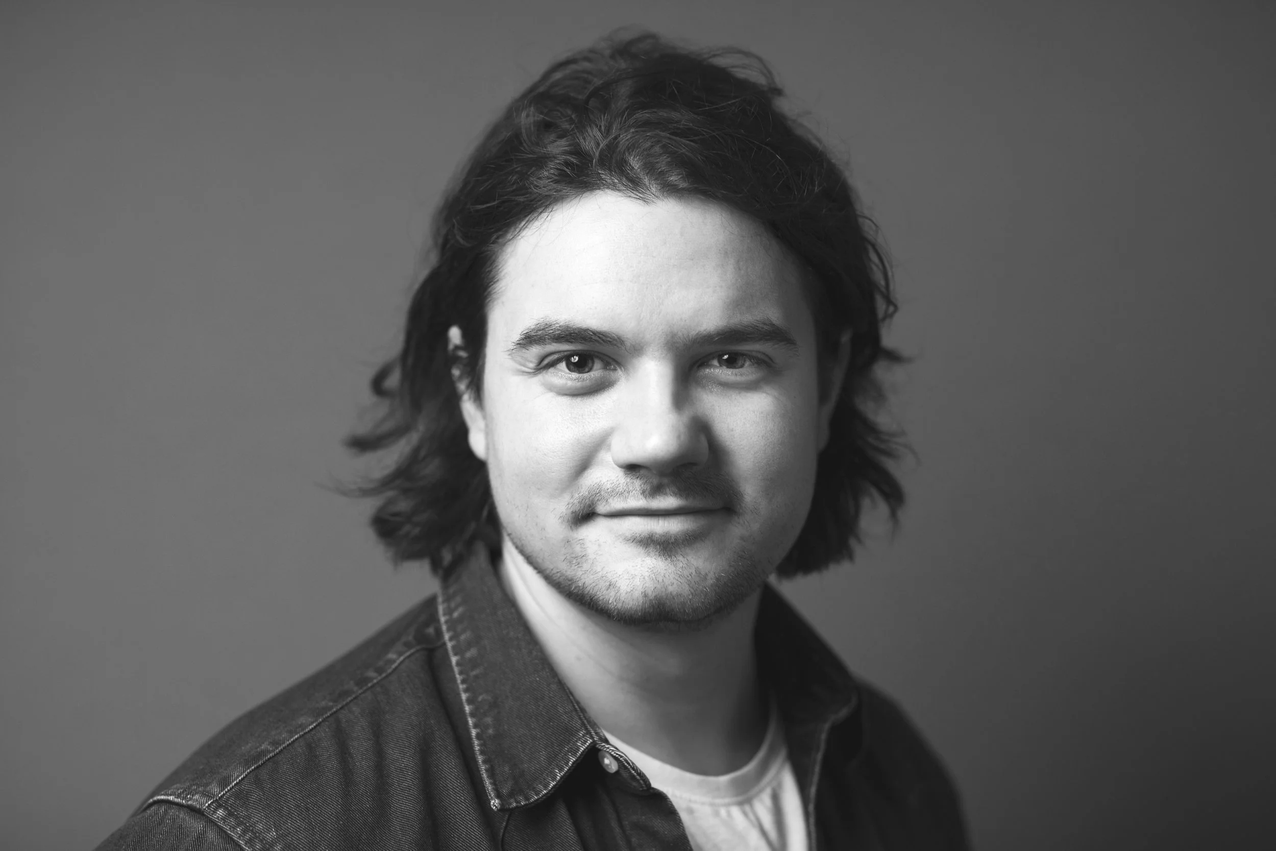 Sort-hvid portrætfoto af en ung mand med mellemlangt, bølget hår, iført en denimjakke og en hvid T-shirt, afbildet mod en neutral baggrund.