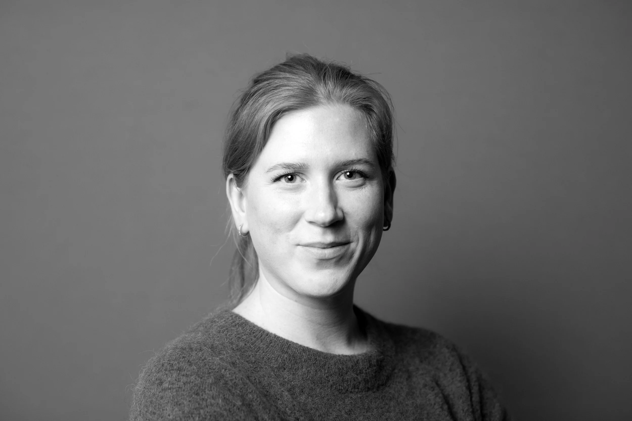 Sort-hvid portræt af en kvinde med let smil, lunget hår, små øreringe, iført en grå strikket sweater, foran en ensfarvet baggrund.