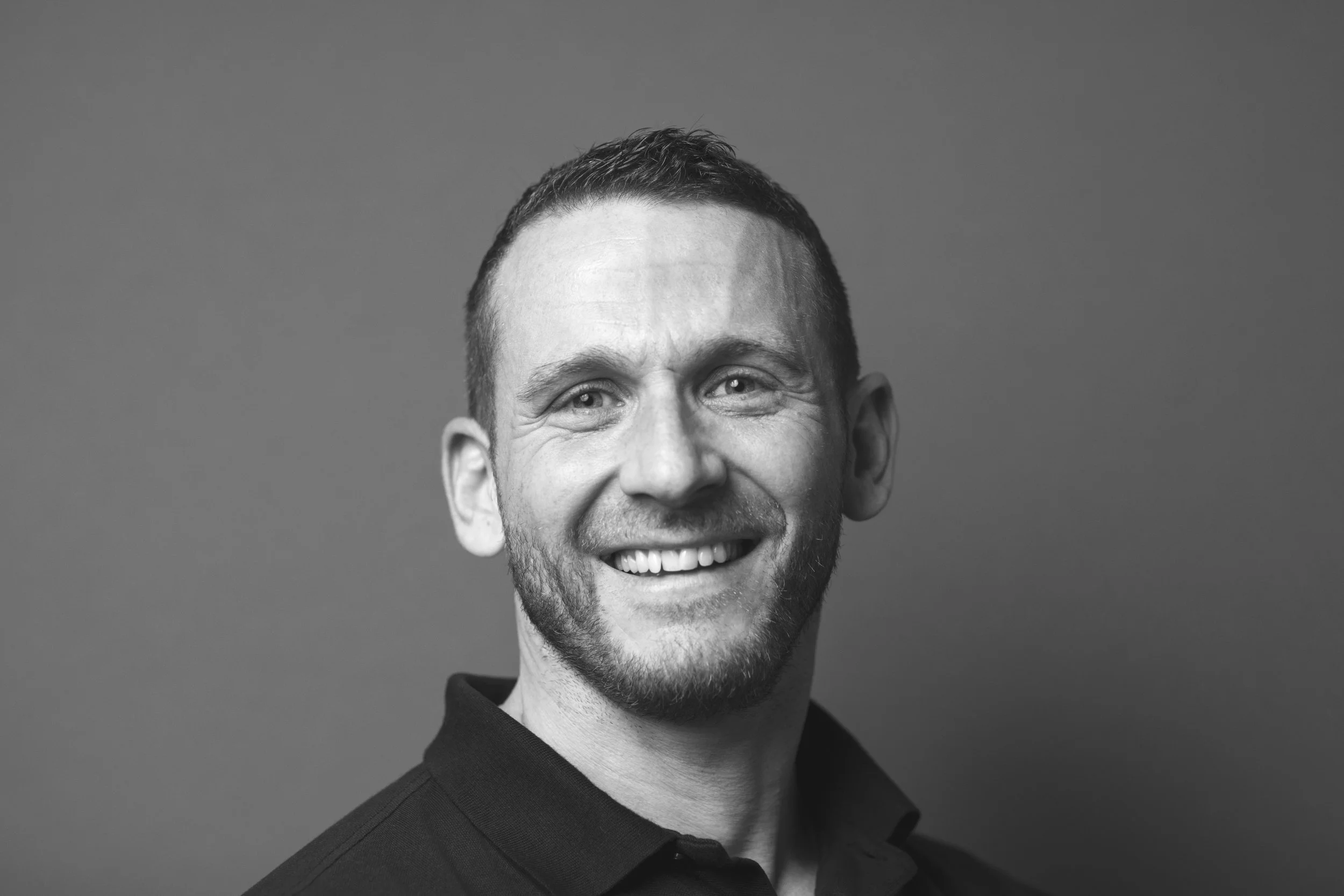 Sort-hvid portrætfotografi af en smilende mand med kort, mørkt hår og skæg, iført sort polo shirt, foran en ensfarvet baggrund.