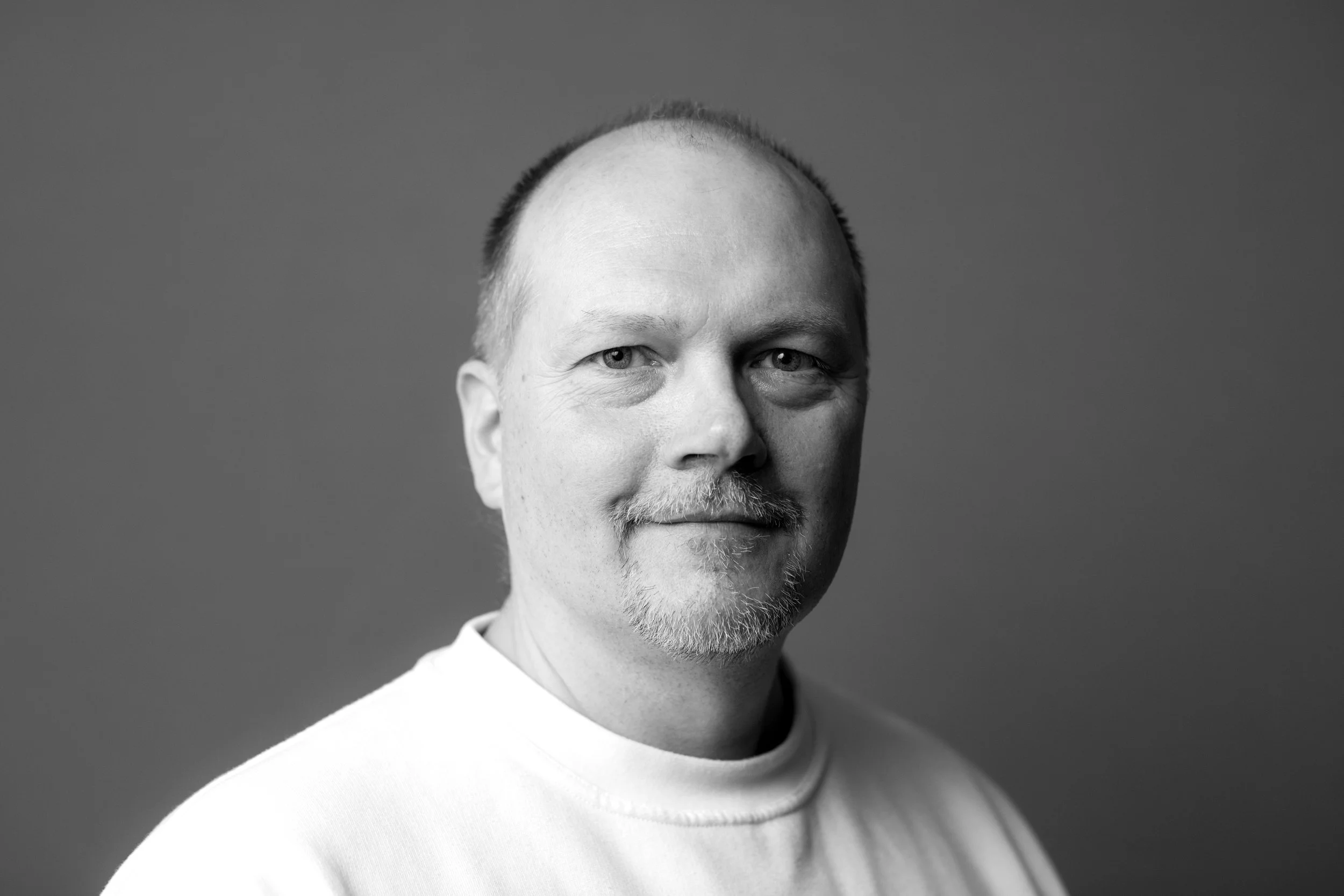 Sort-hvid portrætfoto af en mand med let skæg og blankt hår iført hvid t-shirt, med neutral baggrund.