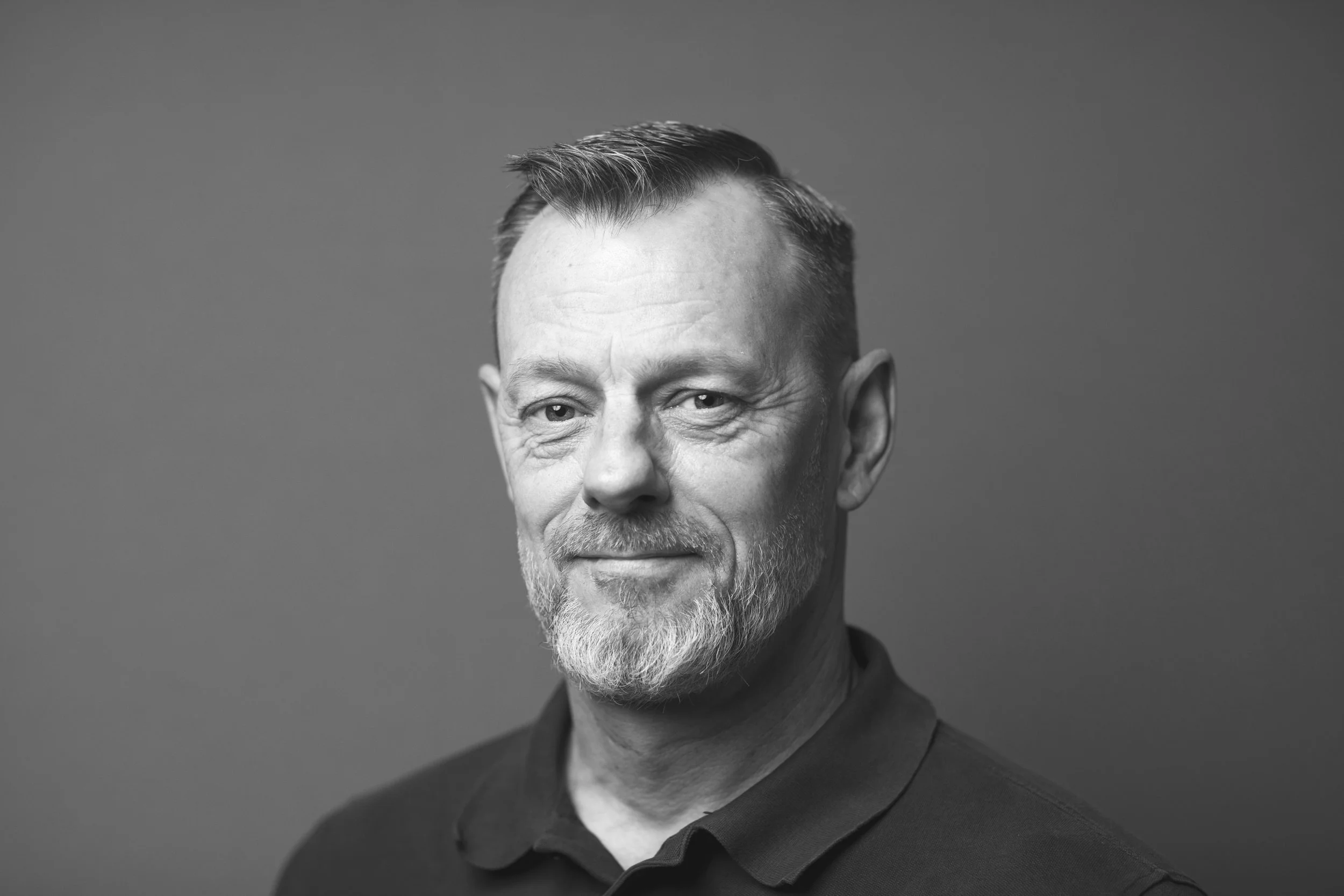 Sort-hvid portræt af en mand med kort, glat, lidt gråt hår og skæg, iført sort skjorte mod en ensfarvet baggrund.