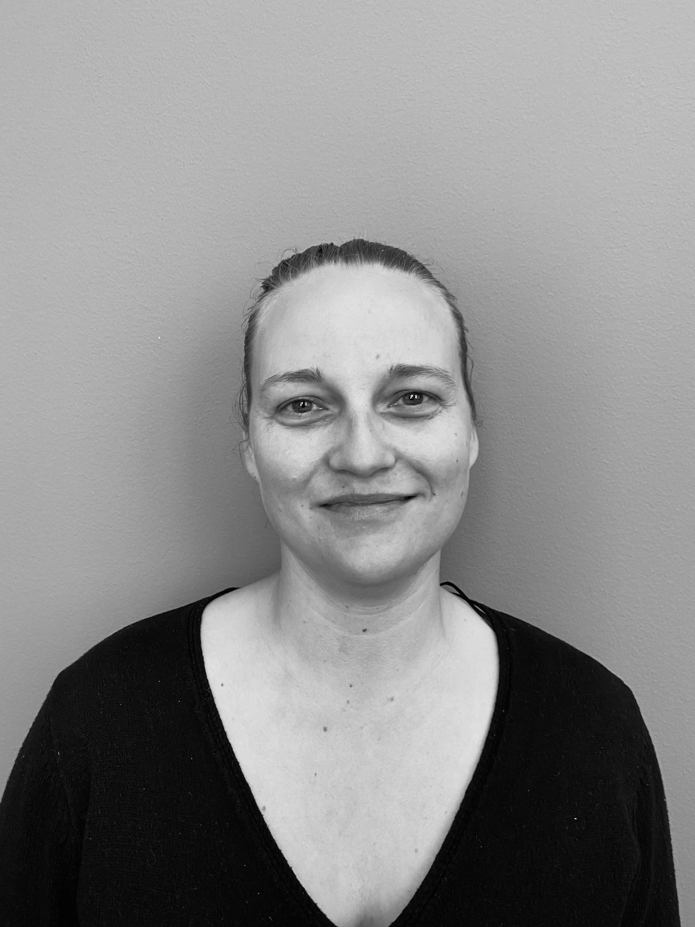 Sort-hvid portrætfotografi af en kvinde med skulderlangt hår, iført sort t-shirt, smilende, foran en glat væg.