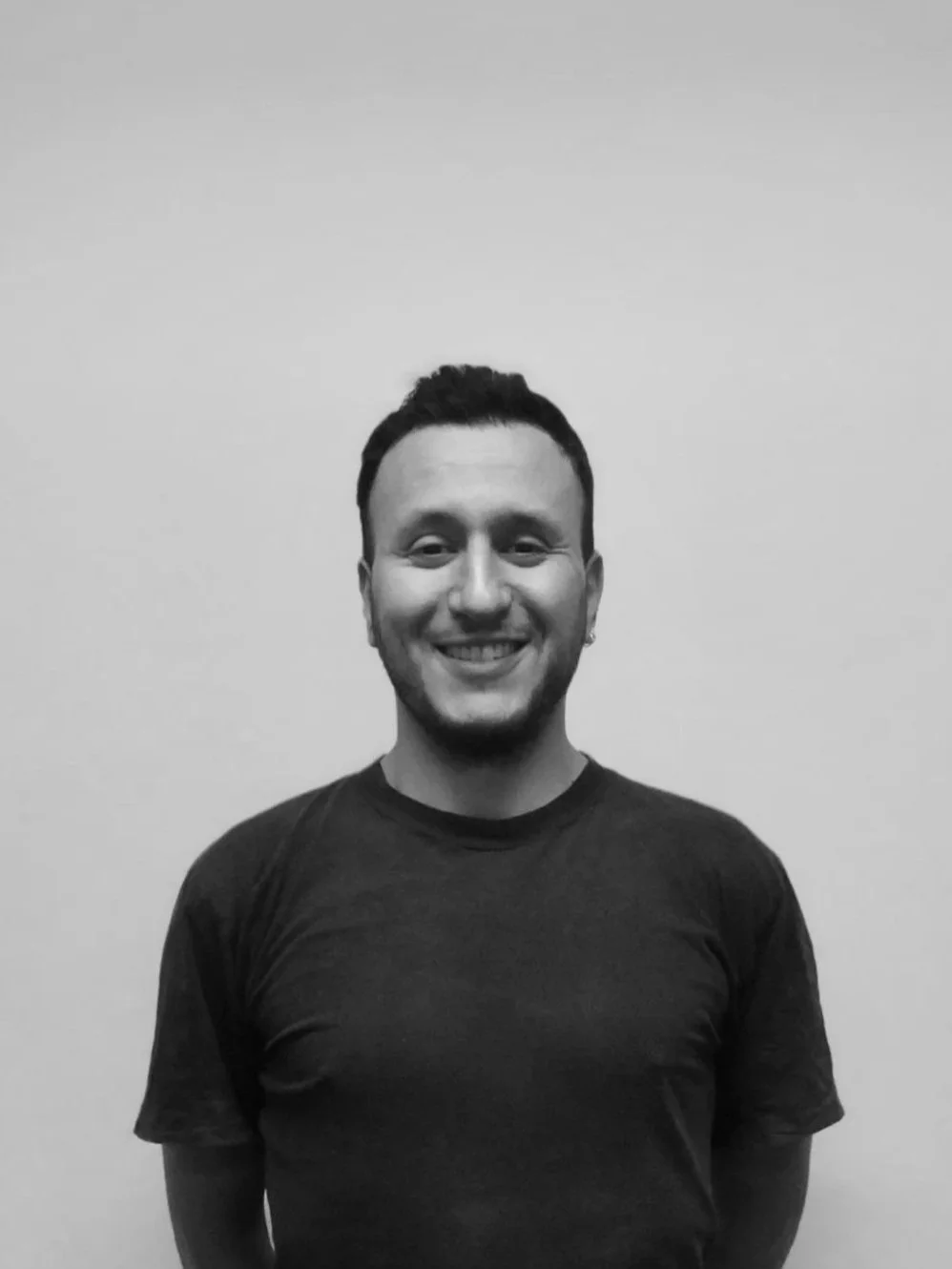 Sort-hvid portrætfotografi af en smilende ung mand med kort hår, iført sort t-shirt, stående mod en neutral baggrund.