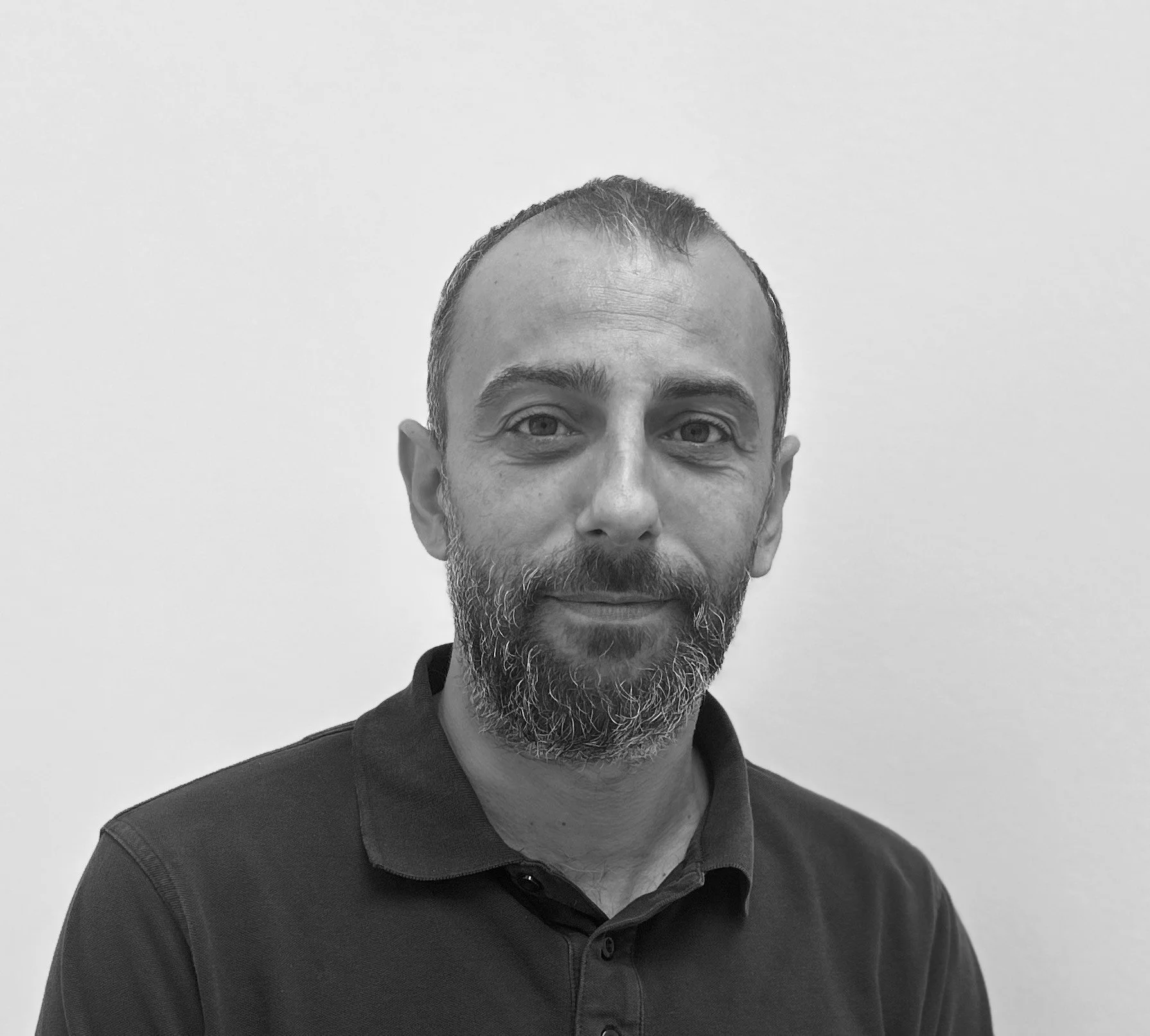 Sort-hvid portrætfoto af en mand med kort, mørkt hår og fuldskæg, iført sort polo-shirt, foran en lys baggrund.