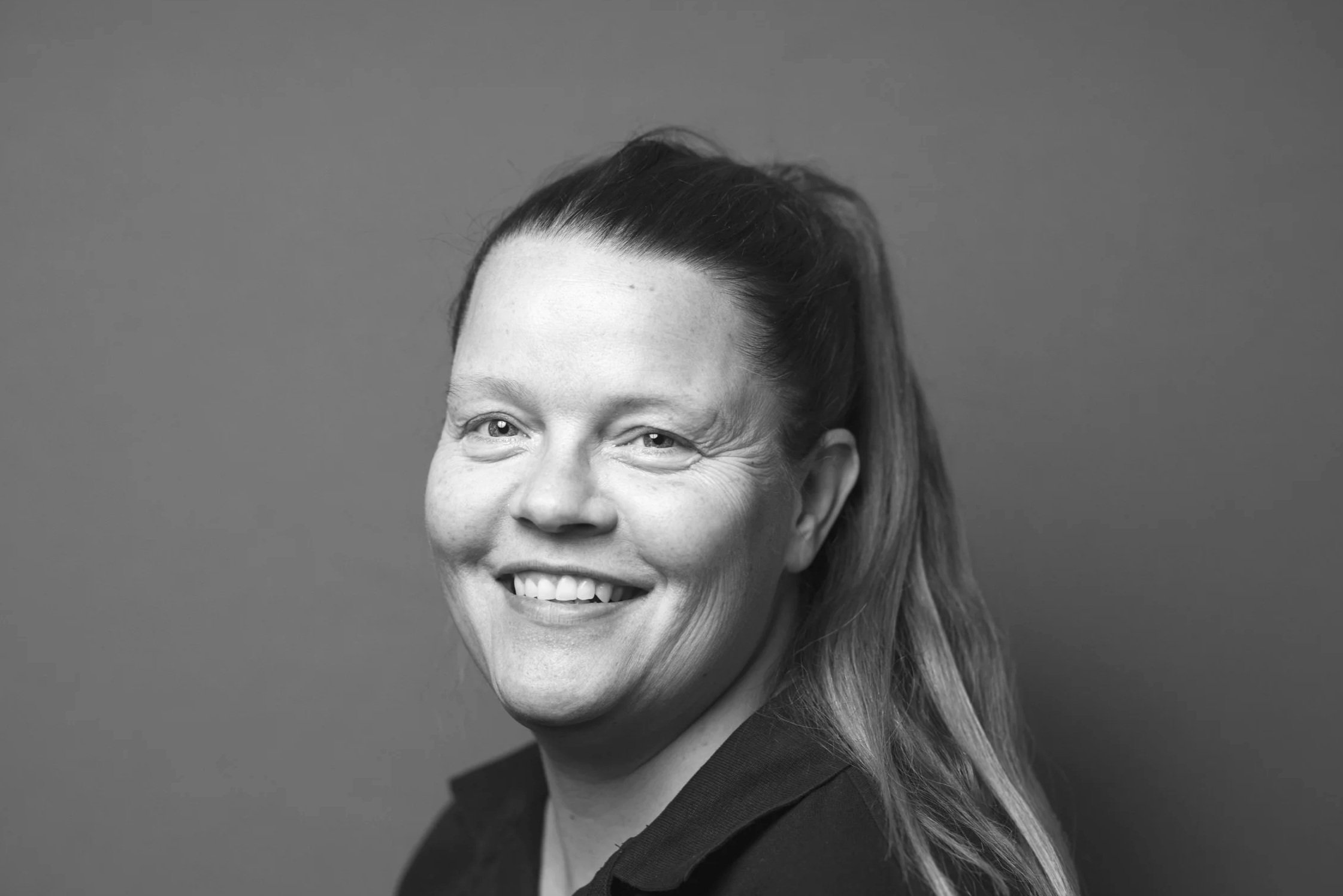 Sort-hvid portræt af en glad kvinde med langt hår, smilende med skæve tænder, iført sort top mod en simpel grå baggrund.