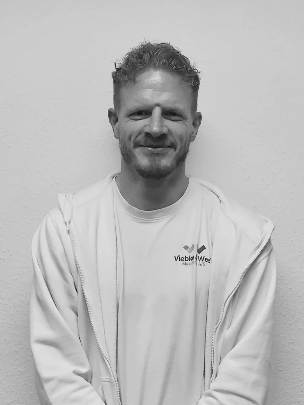 Sort-hvid foto af en mand med kort, krøllet hår, smilende, iført en hvid T-shirt og en hvid hættetrøje med logo, står foran en hvid væg.