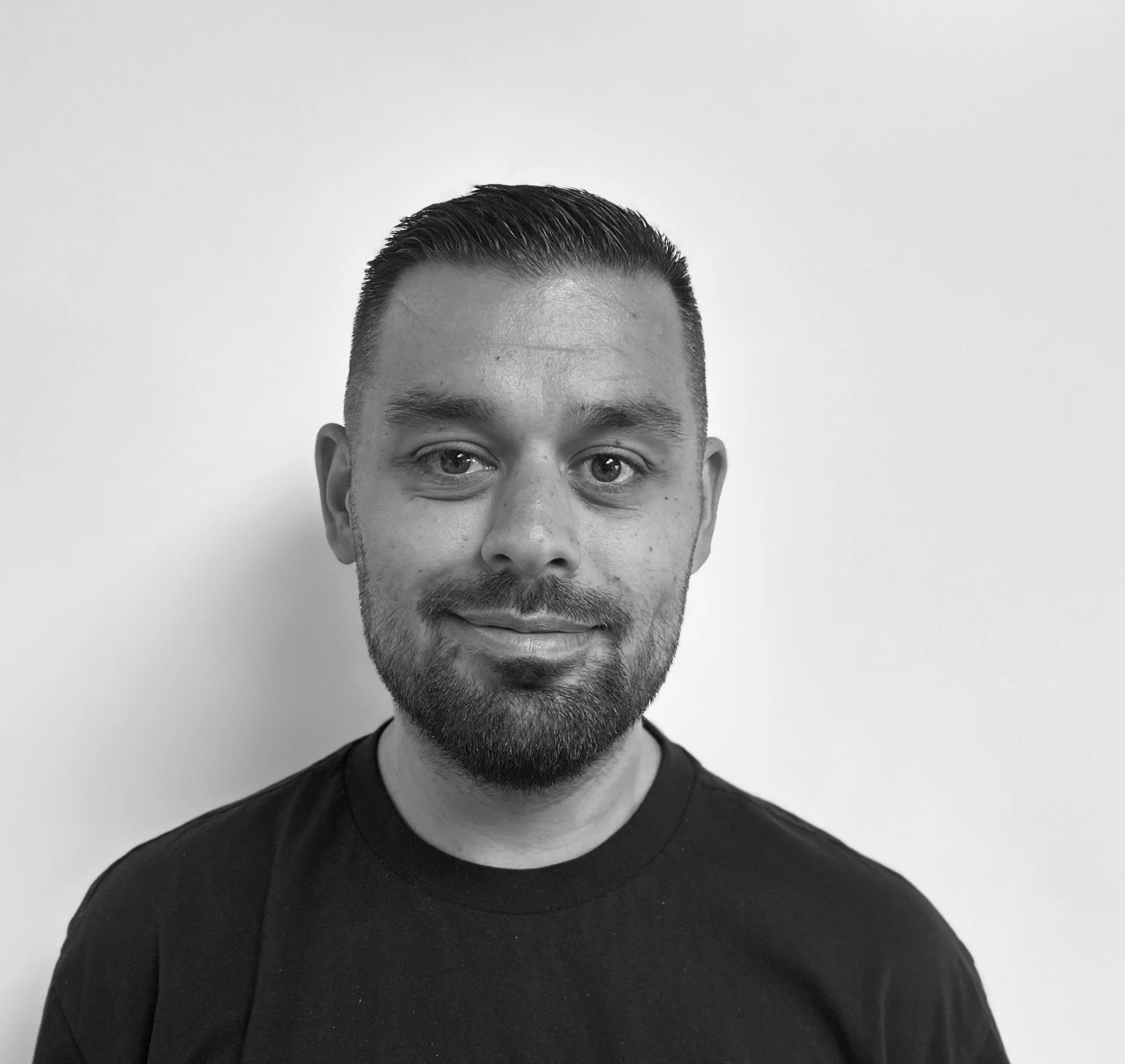 Sort-hvid portrætbillede af en mand med sort T-shirt, kort mørkt hår, fuldskæg, smilende, mod en hvid væg.