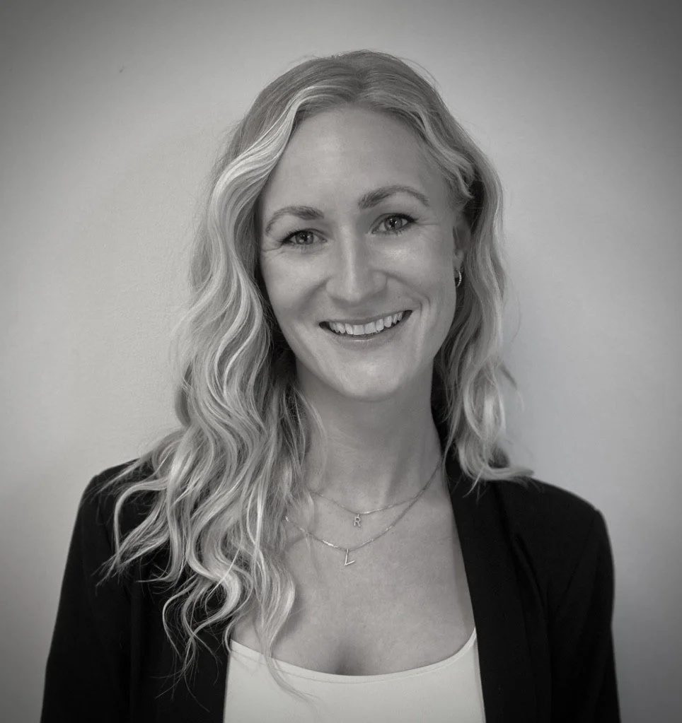 Sort-hvid portrætfotografi af en smilende kvinde med langt, bølget hår, iført en sort jakke og smykker, stående mod en neutral baggrund.