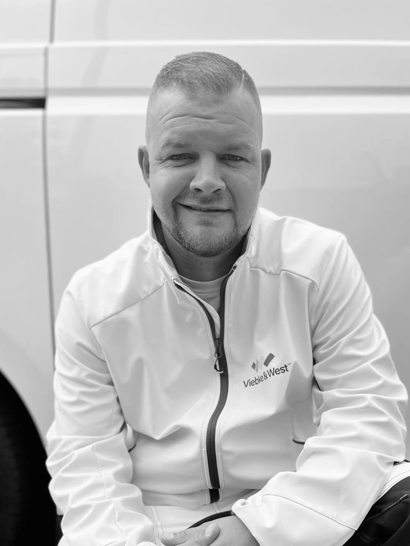 Mand sidder foran en hvid bil, iført hvid jakke med logo 'Viebke & West'