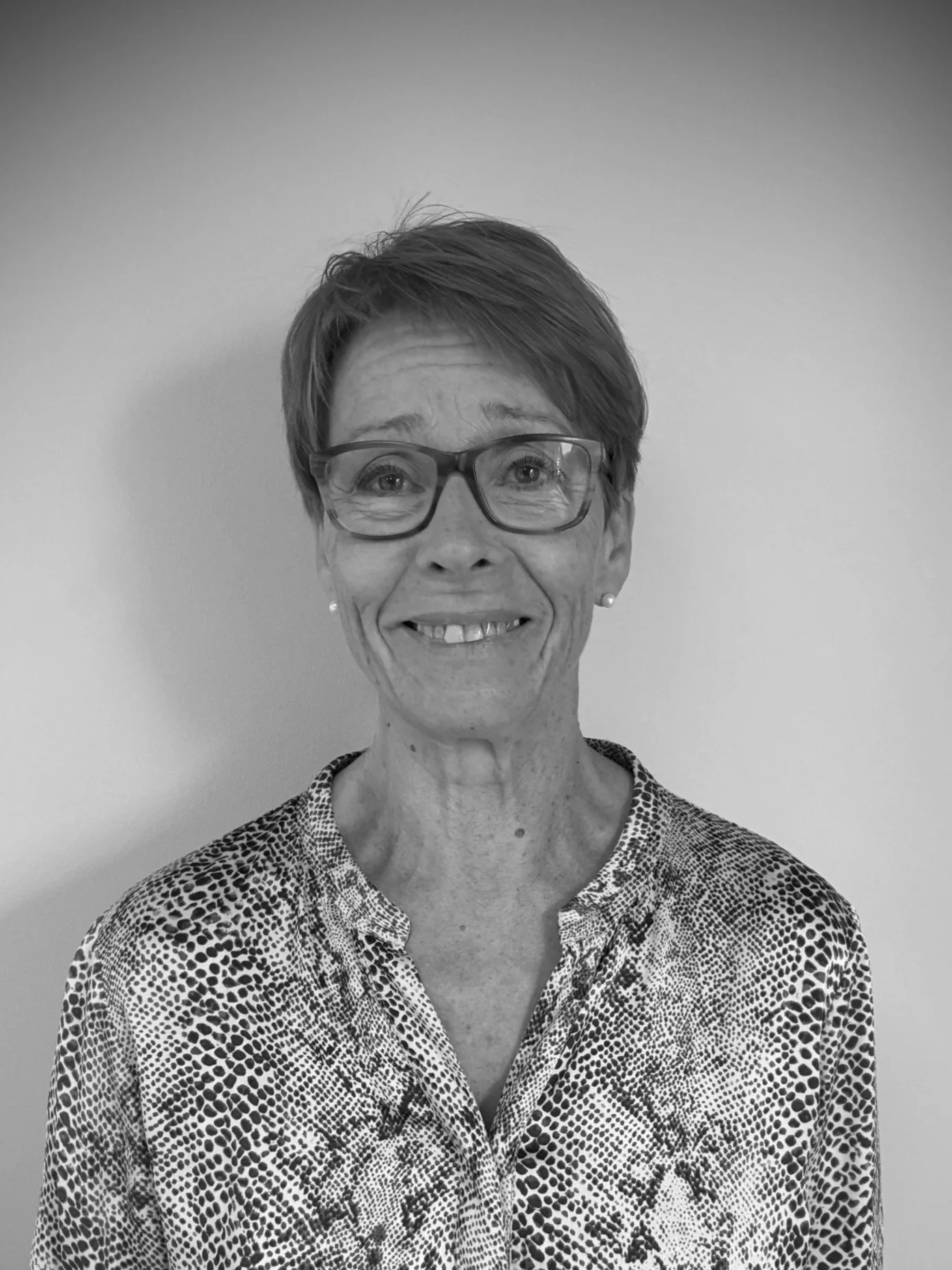 Sort-hvid foto af en smilende ældre kvinde med kort hår, briller og en mønstret skjorte, stående mod en enkel, lys baggrund.
