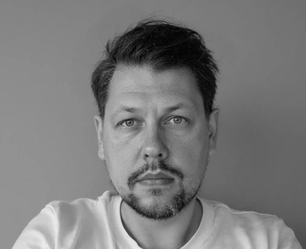 Sort-hvid portræt af en mand med mørkt, glat hår, skæg og skjorte med tern. Han kigger direkte mod kameraet.