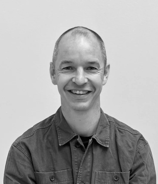 Et sort-hvid fotografi af en mand med kort hår, smilende, iført en jakke.
