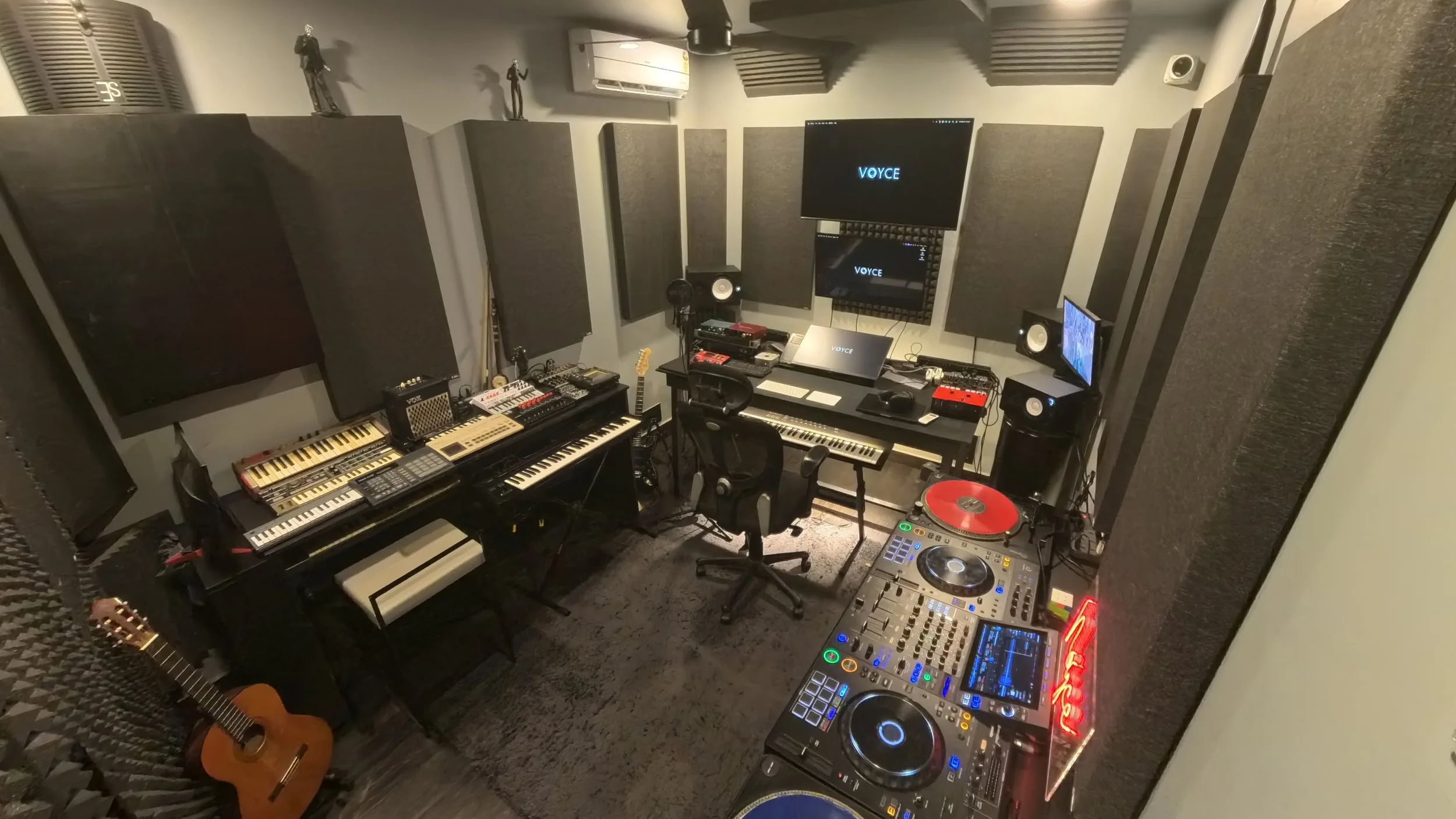 Music production studio with two large monitors displaying digital audio workstation software, surround sound speakers, musical instruments, and soundproof walls.