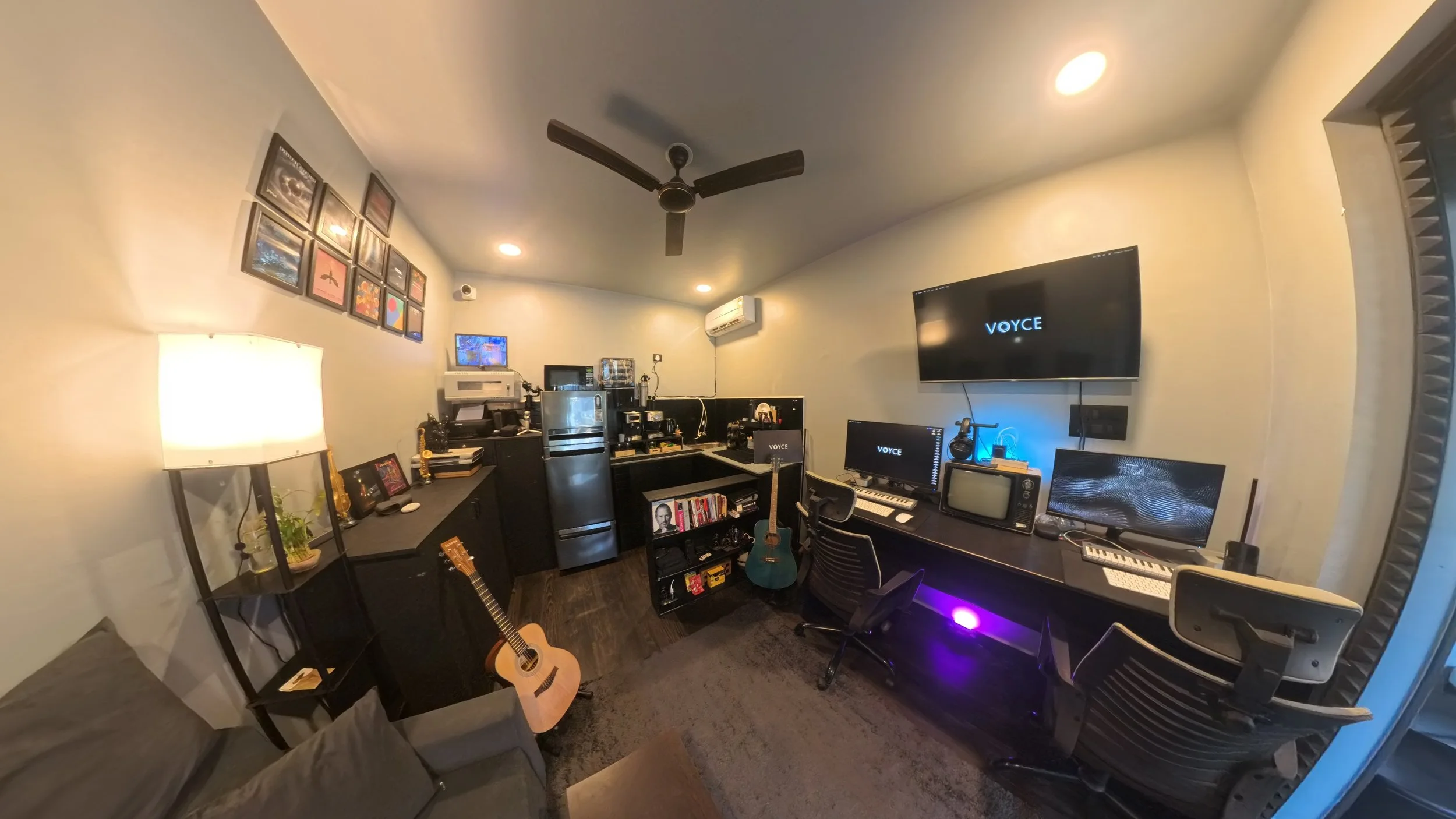 A music studio workspace with a keyboard, two laptops, speakers, a desktop monitor, and a camera on a black desk, with a window showing an outdoor balcony and greenery.