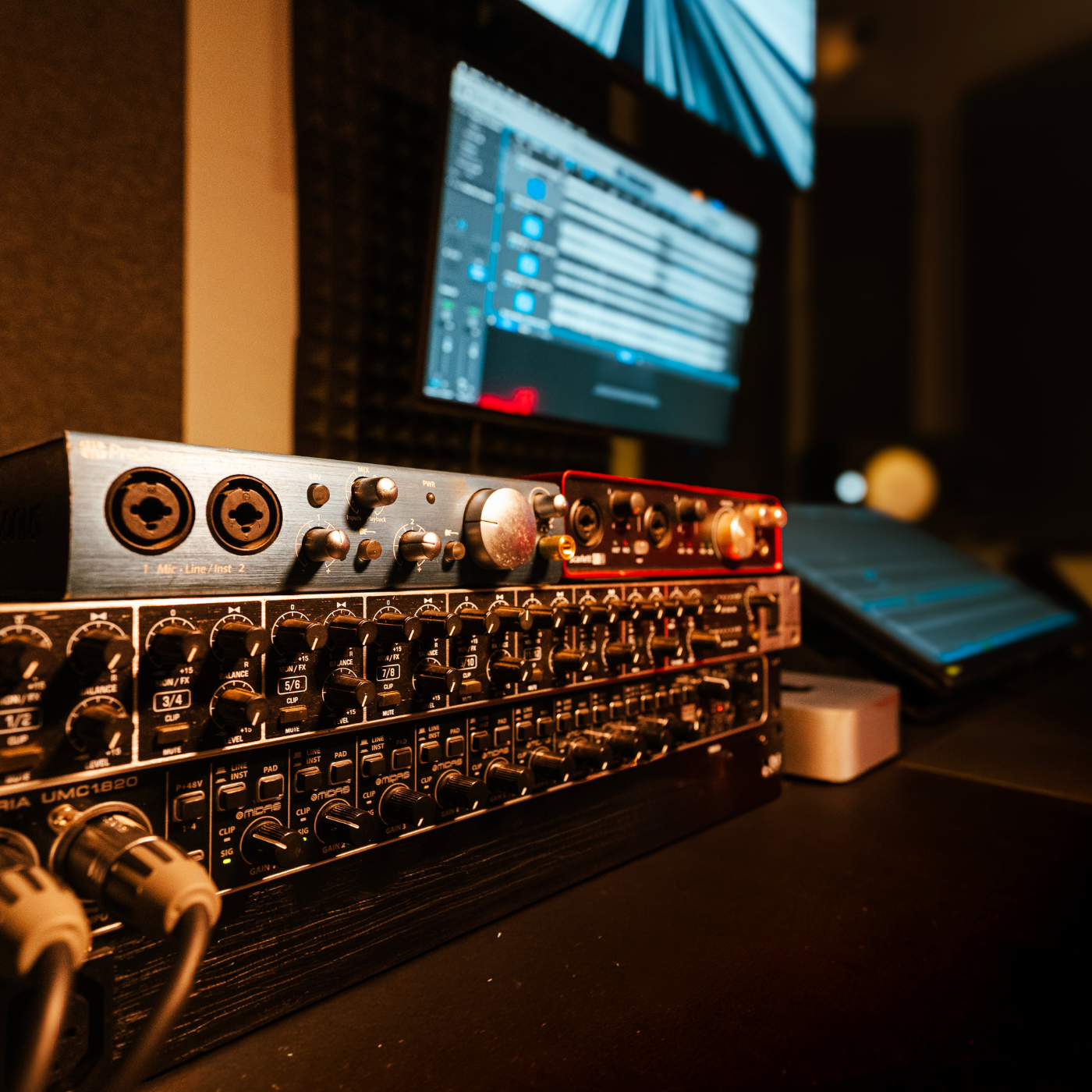 Close-up of a professional audio mixing console with various knobs and sliders, computer screen in the background displaying digital audio workstation software, and studio foam panels on the wall.