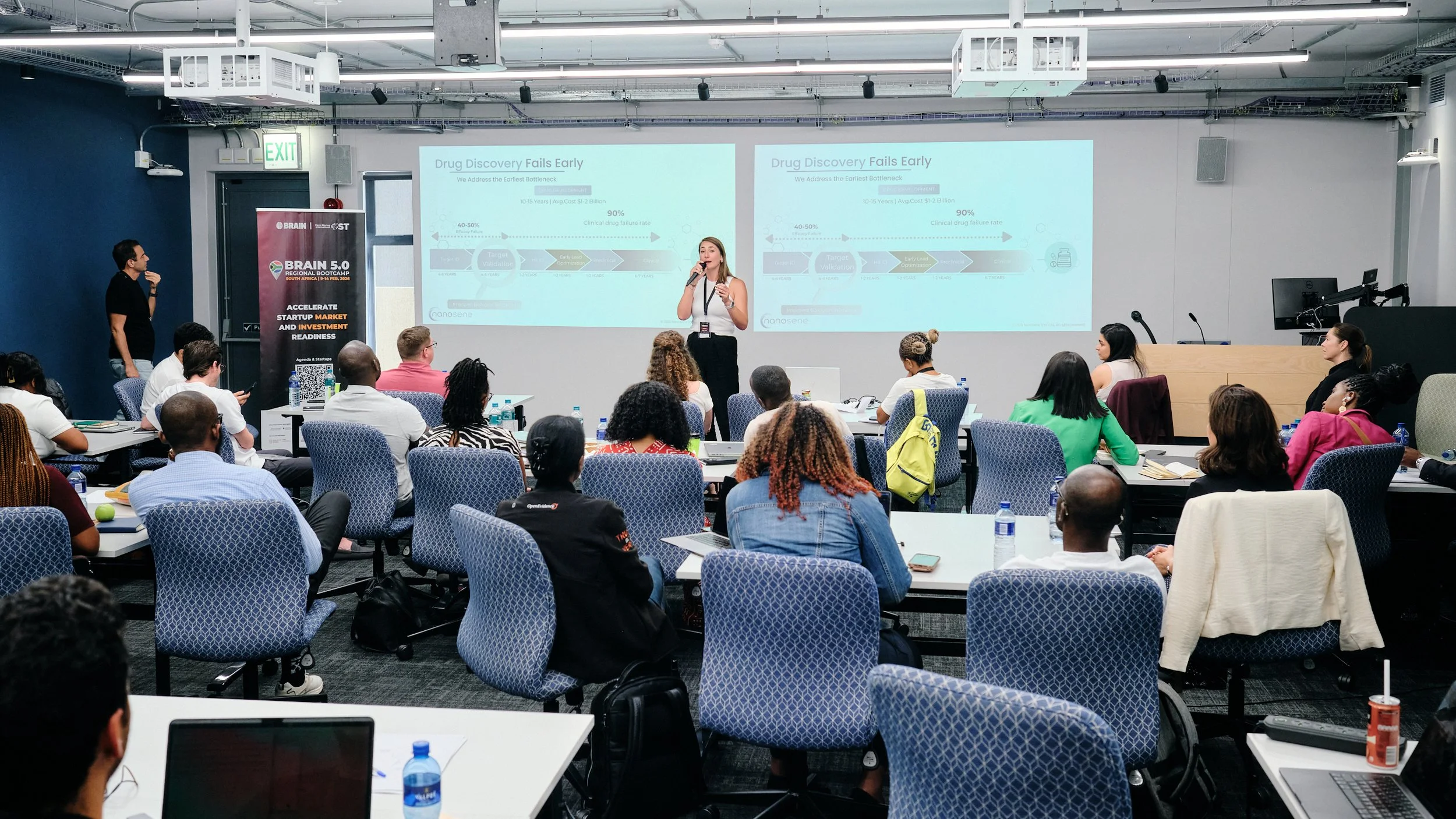 A woman faces an audience, speaking into the mic. The audience is seated with backs to the camera. Behind the woman is a powerpoint presentation.