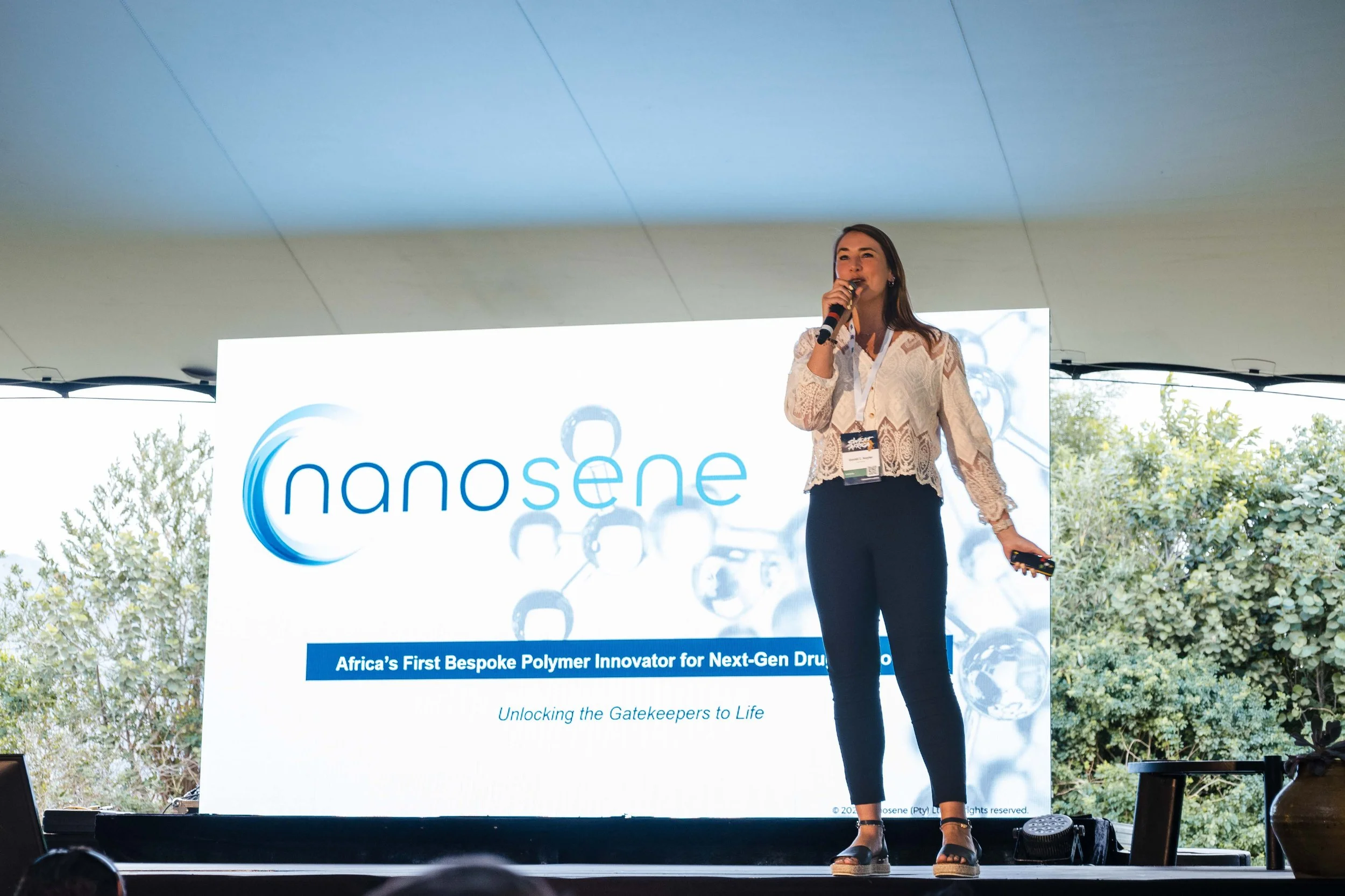 A ceo stands on a stage in front of a powerpoint with bushes in the background. She is speaking into a microphone. The slide contains the Nanosene logo, as well as slogans partially hidden behind the CEO.