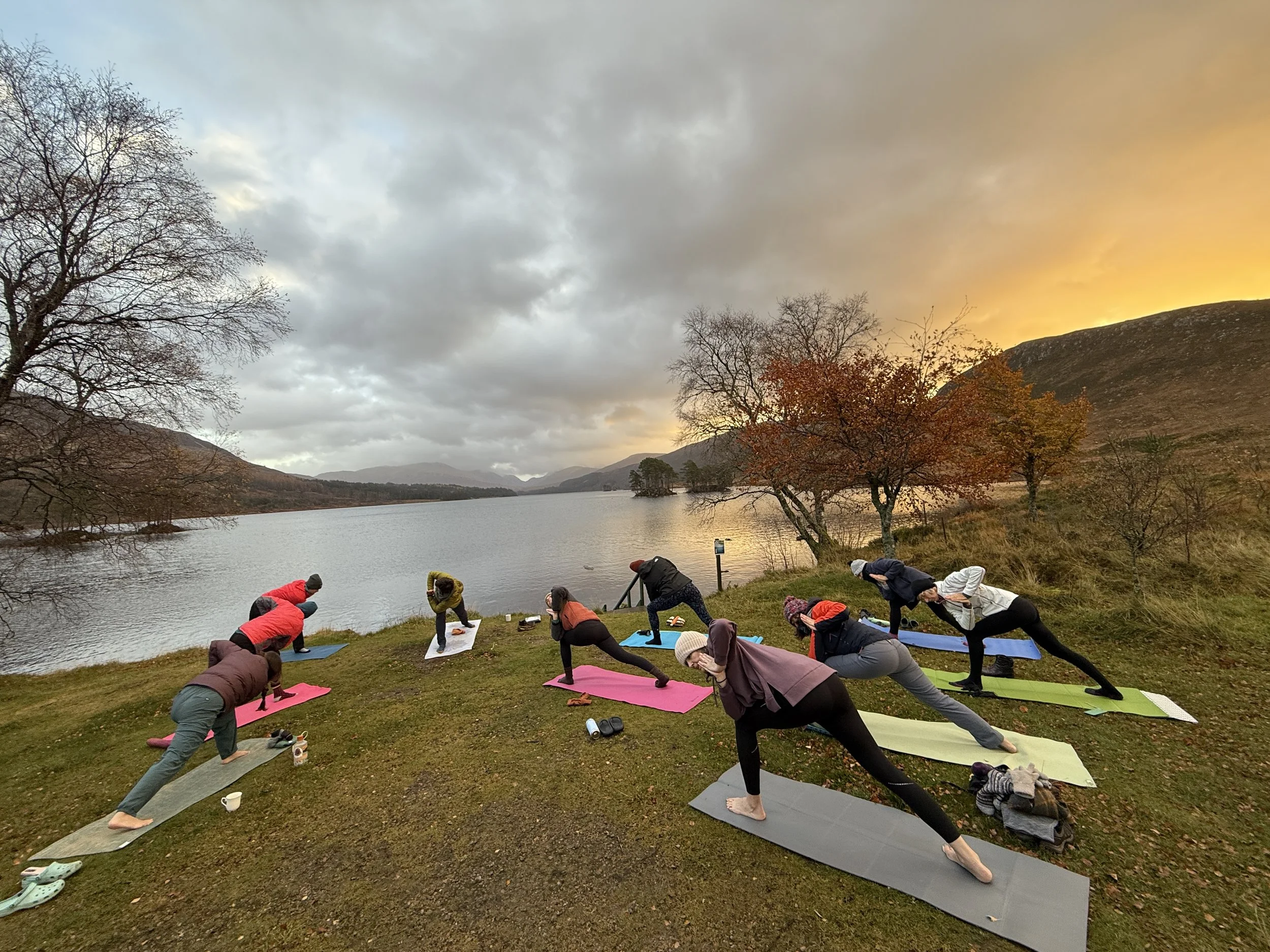 Group of people practicing yoga outdoors on colorful mats during sunset by a lake with trees and mountains in the background.