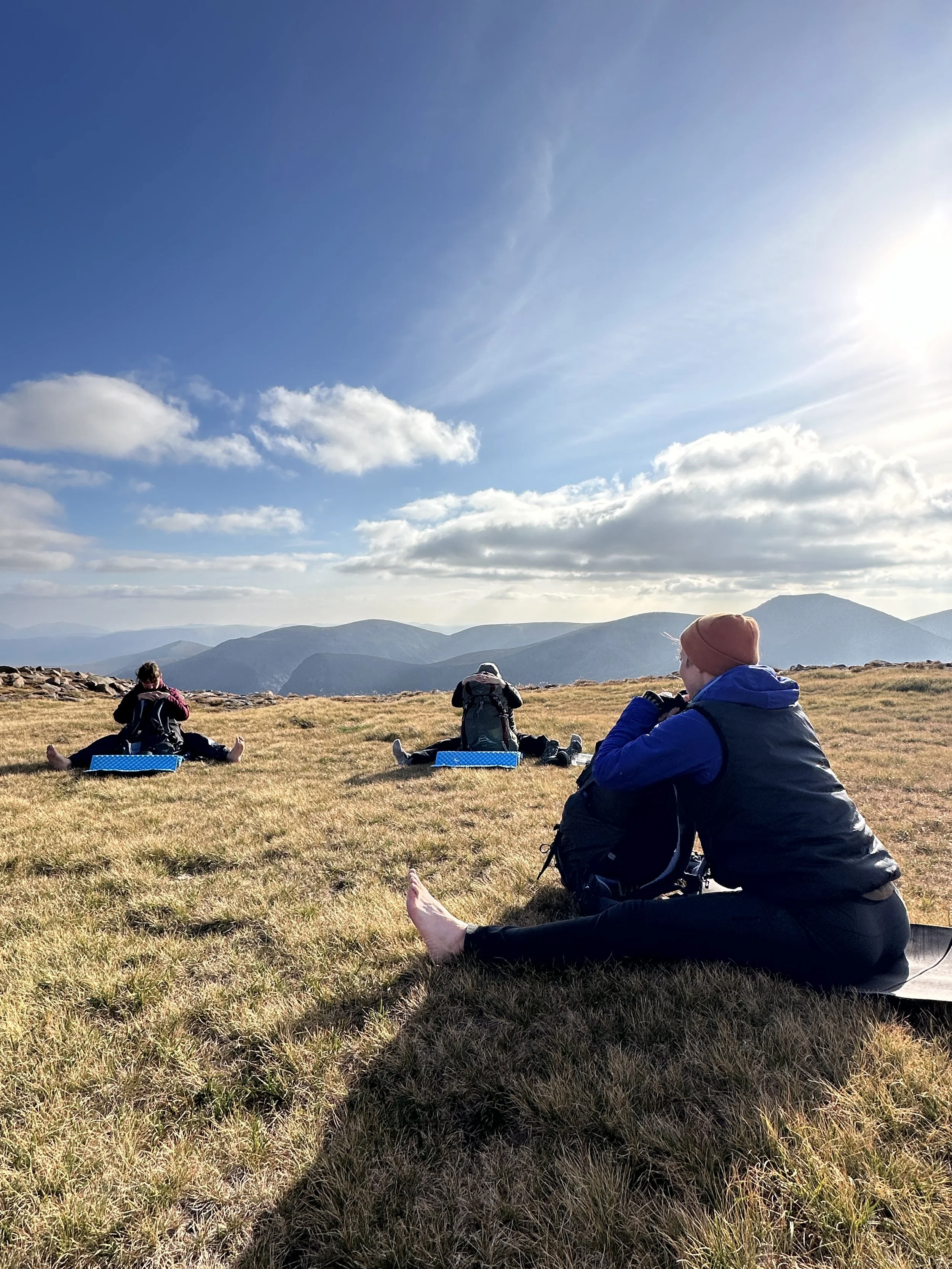 Three hikers resting on a grassy hill with mountains in the background, sitting on blue mats under a partly cloudy sky during daytime.