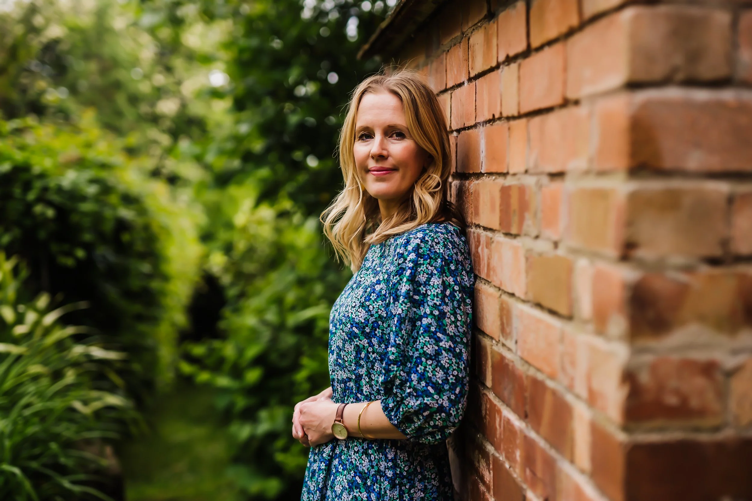 A woman with wavy blonde hair stands next to a brick wall outdoors, with green foliage in the background, wearing a blue floral dress and a wristwatch, smiling softly.