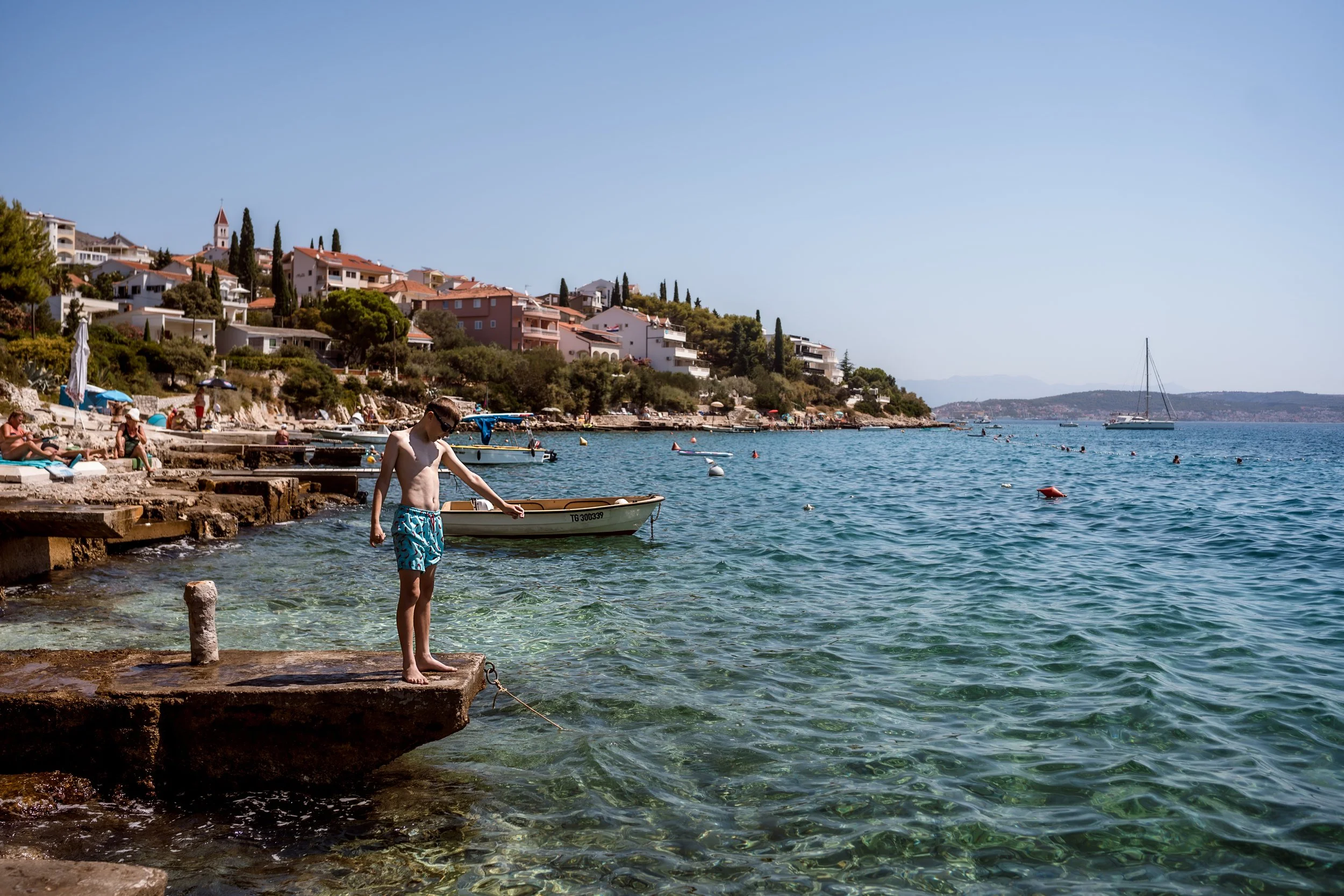 Boy standing on a concrete dock at the edge of a clear blue water seaside, with a small boat and houses on a hillside in the background.