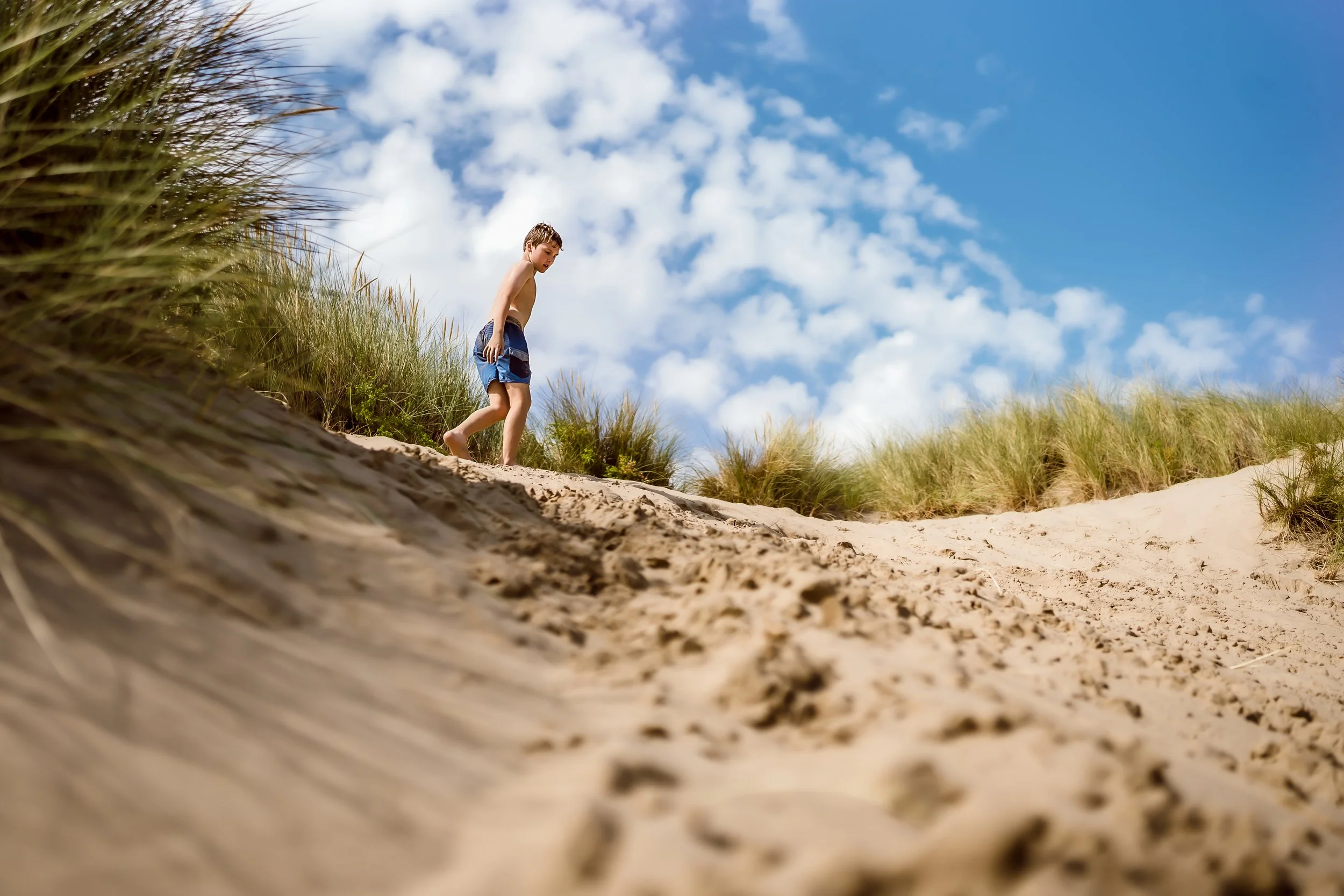 Young boy in swim trunks walking along a sandy beach with grass-covered dunes under a blue sky with scattered clouds.