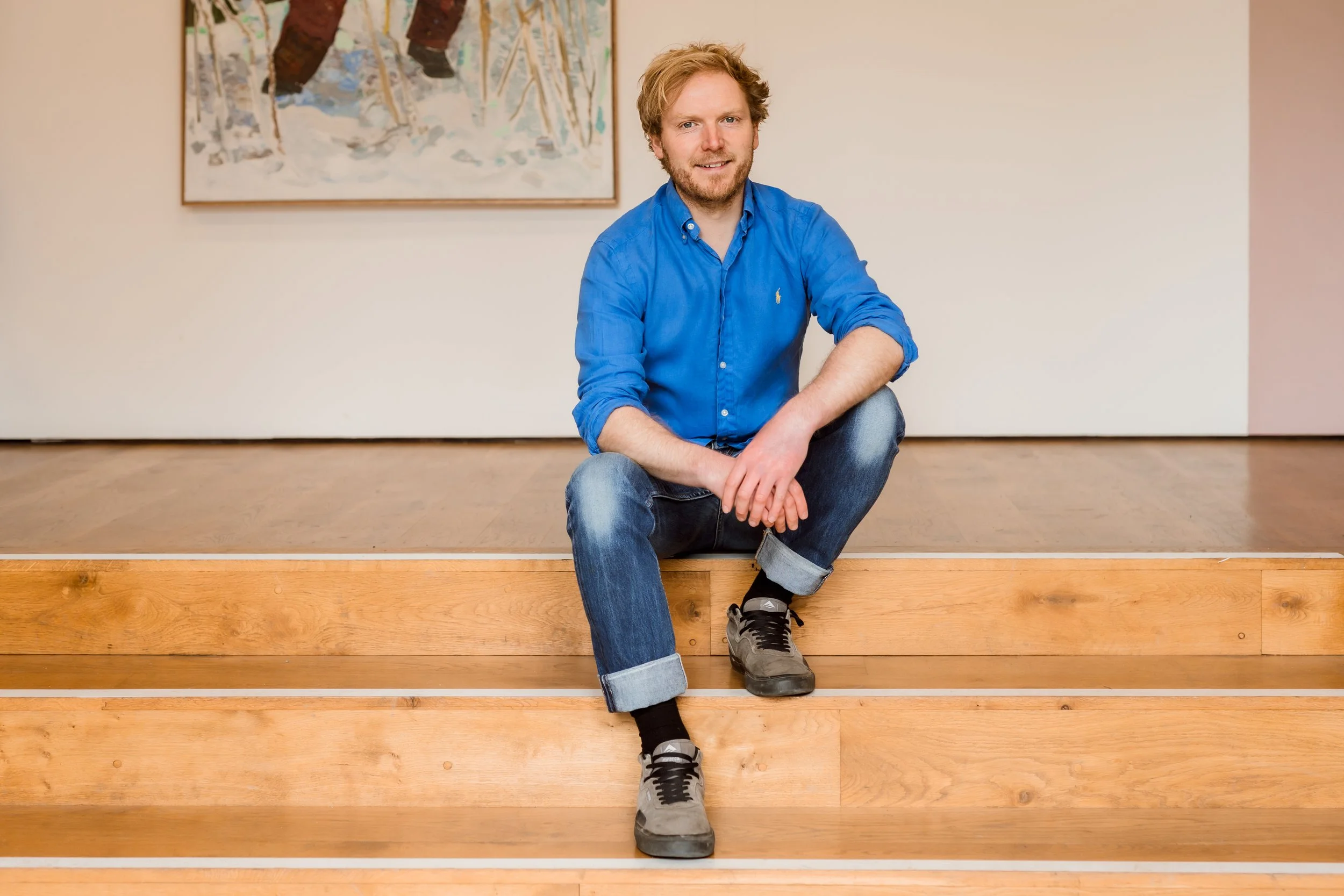 A man with curly blonde hair and a beard, wearing a blue shirt and jeans, is squatting on a wooden step in front of a white wall with a colorful painting behind him. He is smiling at the camera.