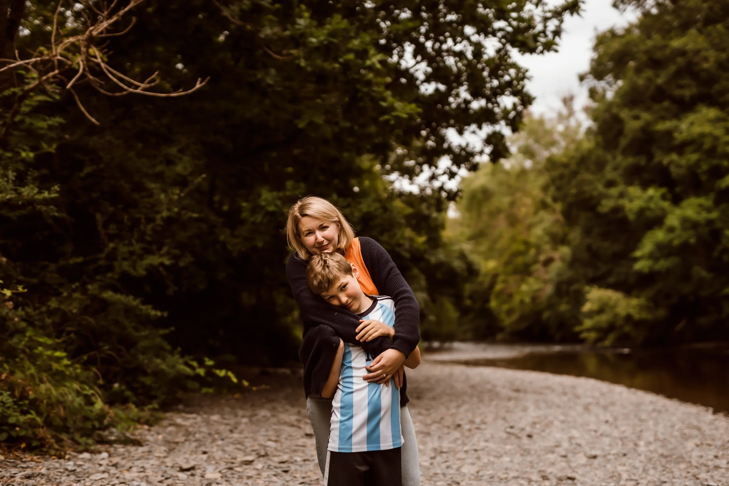 A woman and a young boy are standing together outdoors on a gravel path, with the woman embracing the boy from behind. They are surrounded by dense green trees and overcast skies.