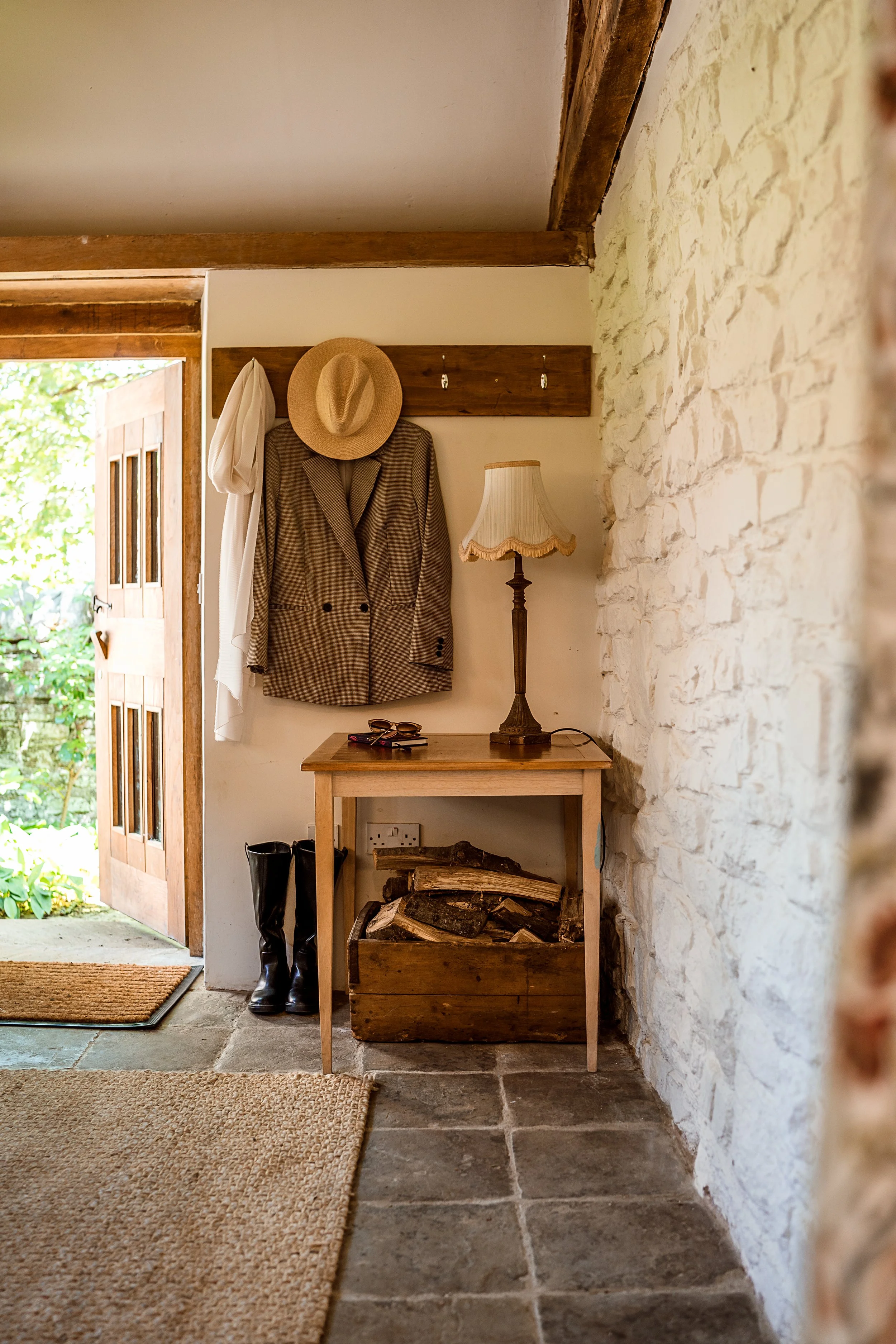 Entryway with wooden door open to green outdoor garden, coat rack with plaid blazer, straw hat, and white cloth, vintage lamp on small wooden table, pair of black rain boots, and firewood in a wooden box under the table, stone and painted walls, tiled floor, at home.