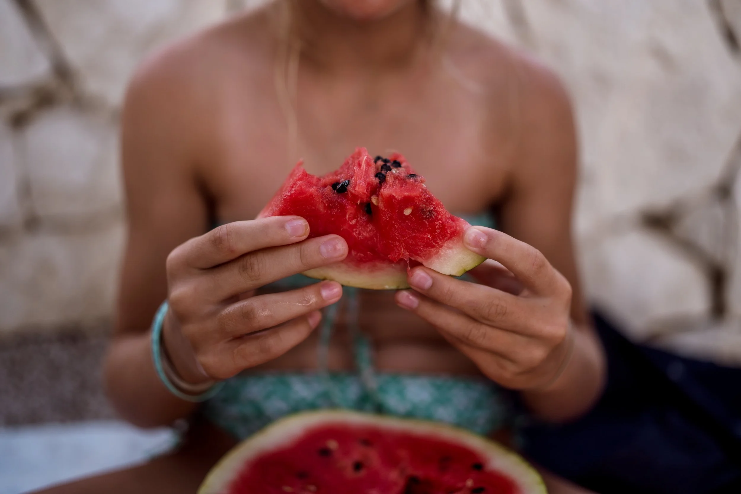A person holding a slice of watermelon with black seeds, with more watermelon slices on a table in front.