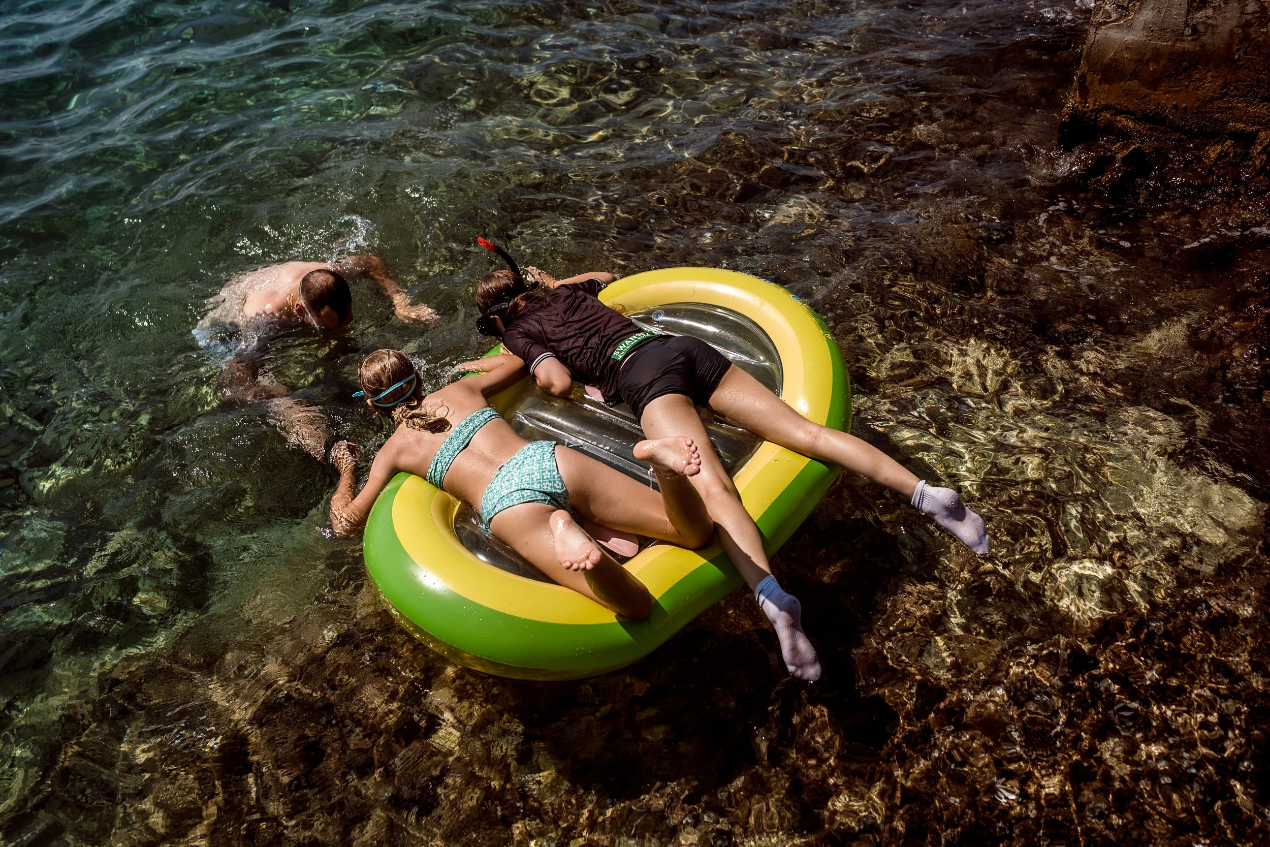 Three children in swimsuits playing in a small inflatable boat in shallow water near a rocky shoreline.