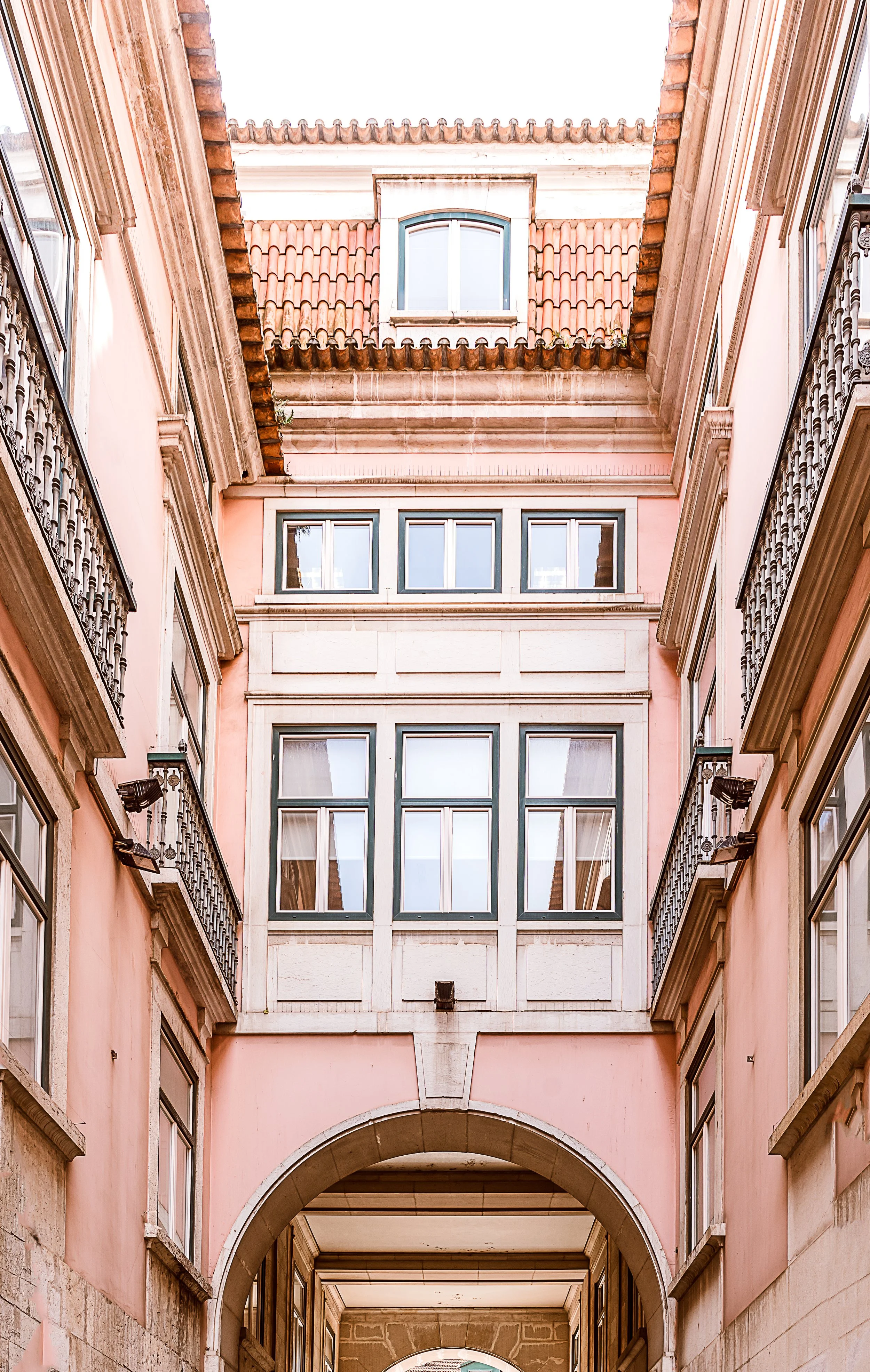 A narrow courtyard surrounded by pink building facades with arched hallways, windows, and balconies with black wrought iron railings, leading to a view of the sky.