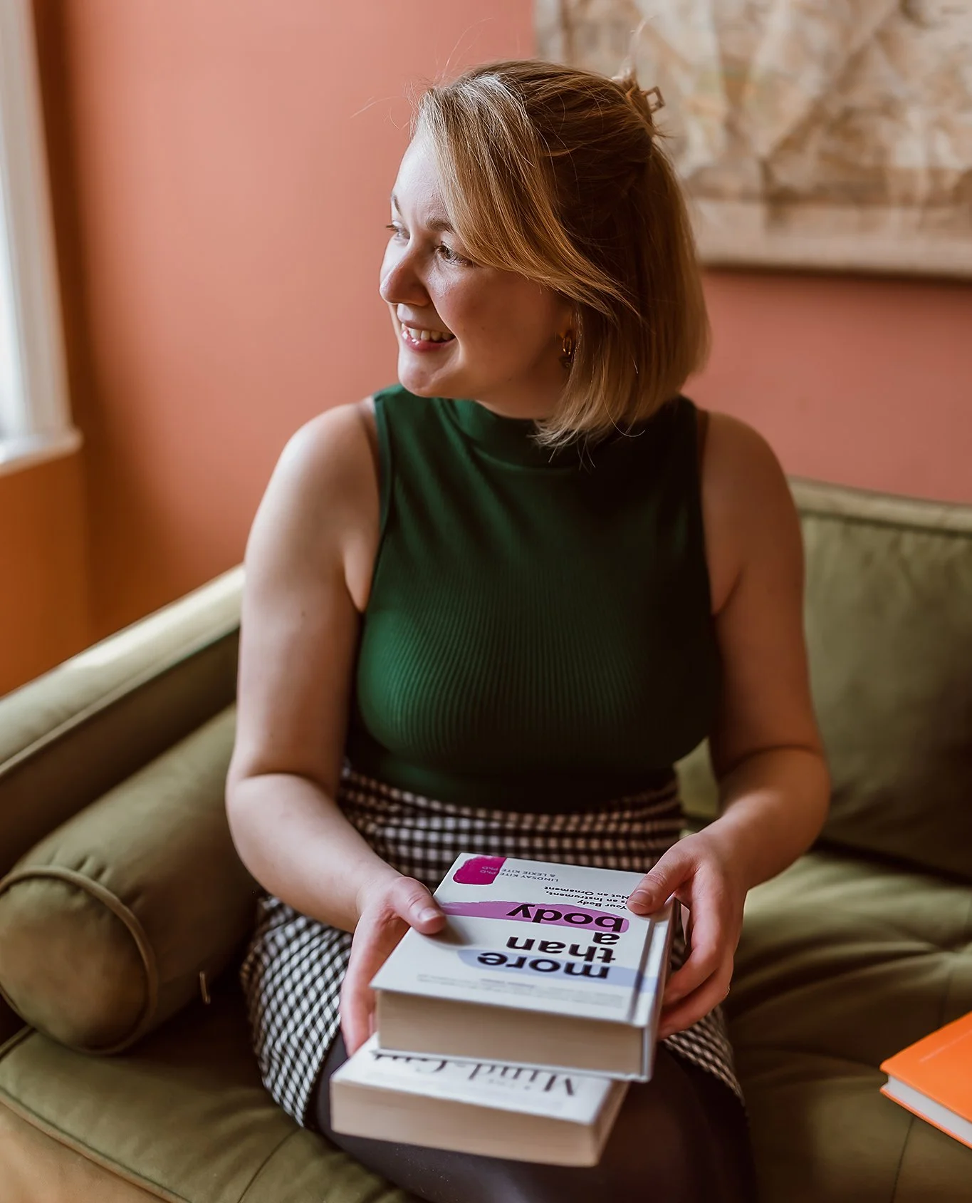 A woman with shoulder-length reddish hair smiling while sitting on a green cushioned bench, holding a book titled 'More Than a Boy' and a small box, in a room with pink walls and artwork in the background.