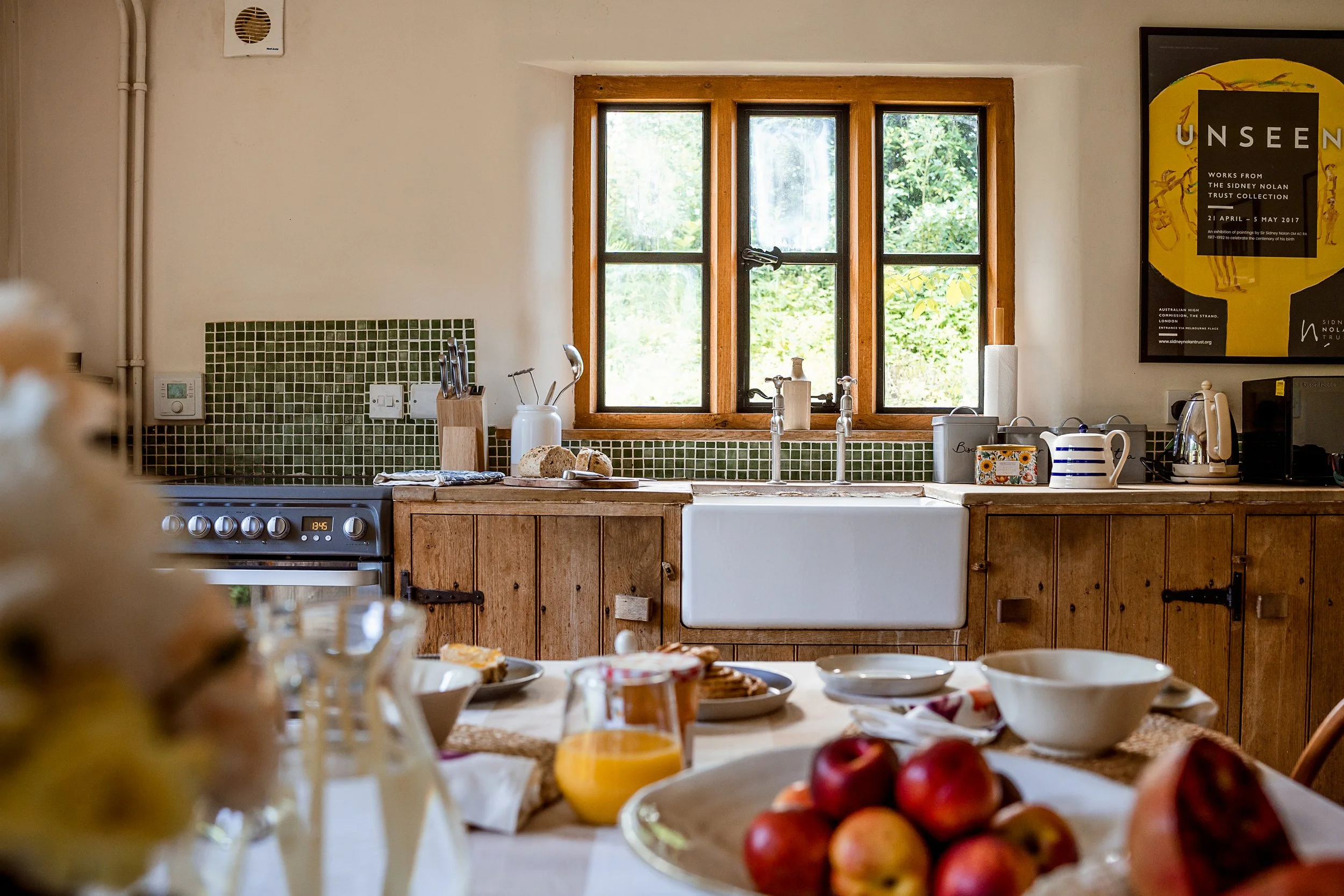 A kitchen with a large window, green tile backsplash, and wooden cabinetry. The countertop holds a loaf of bread, utensils, and a white ceramic pitcher near a sink. To the right, there are appliances and a colorful poster on the wall. In the foreground, a table is set with plates, bowls, glasses, and a plate of apples.
