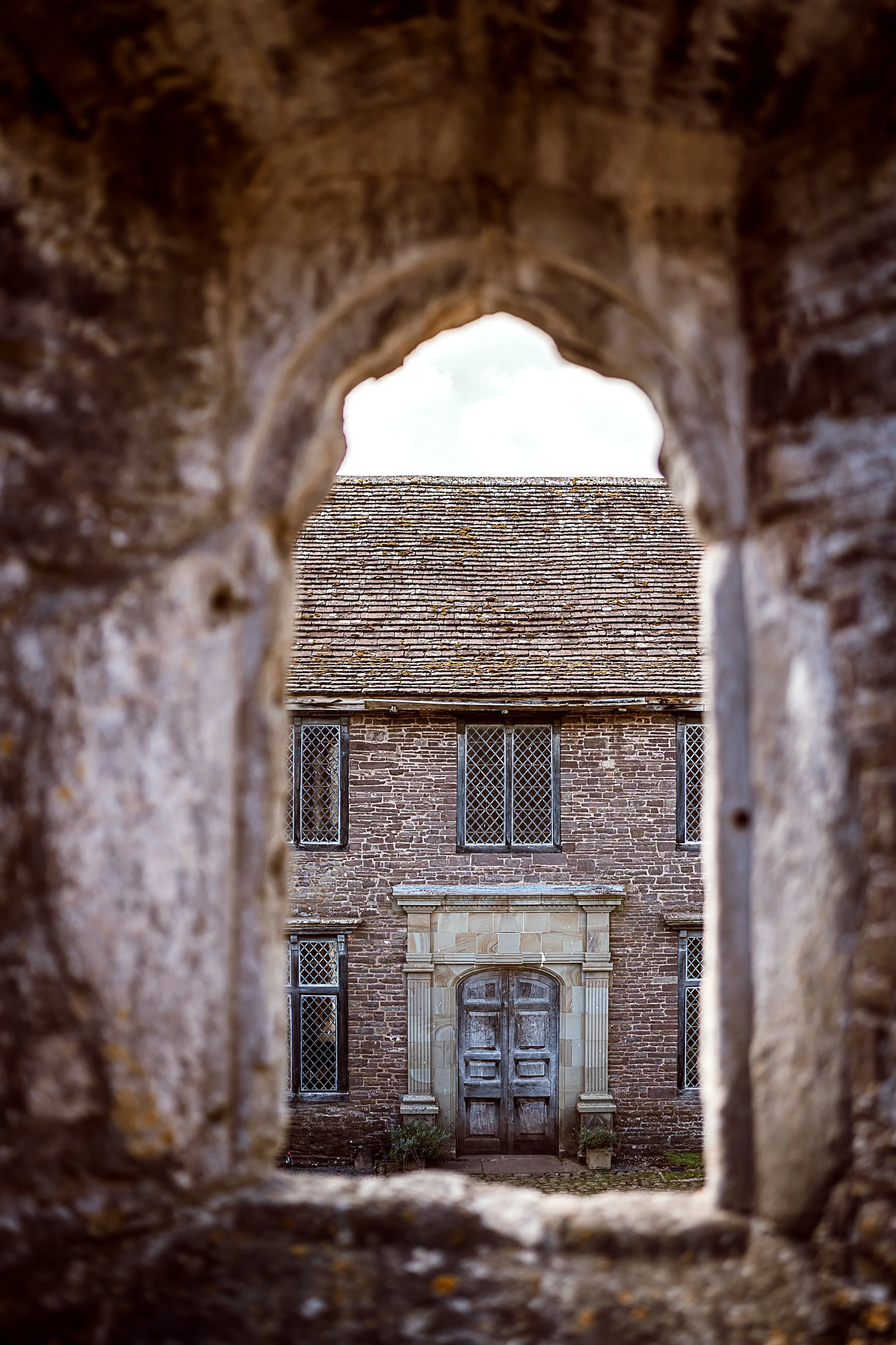 View of a brick building with a large wooden door, seen through a stone archway with a weathered appearance, and lattice windows above.