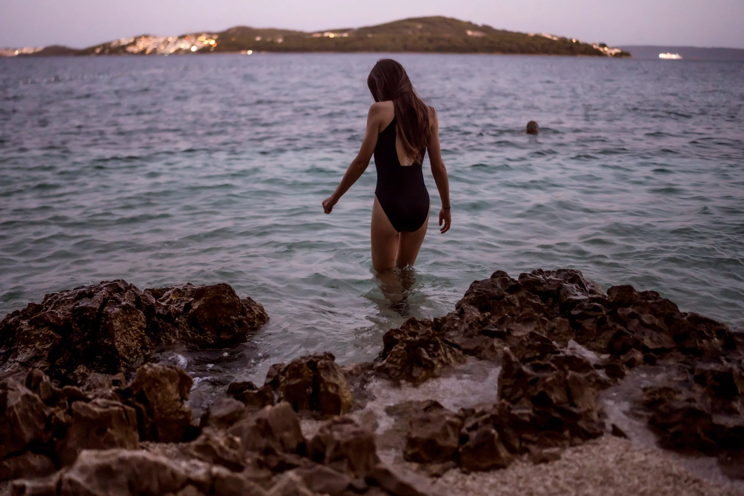 A woman in a black swimsuit walks into the water near rocky shoreline at dusk, with an island and a boat in the background.