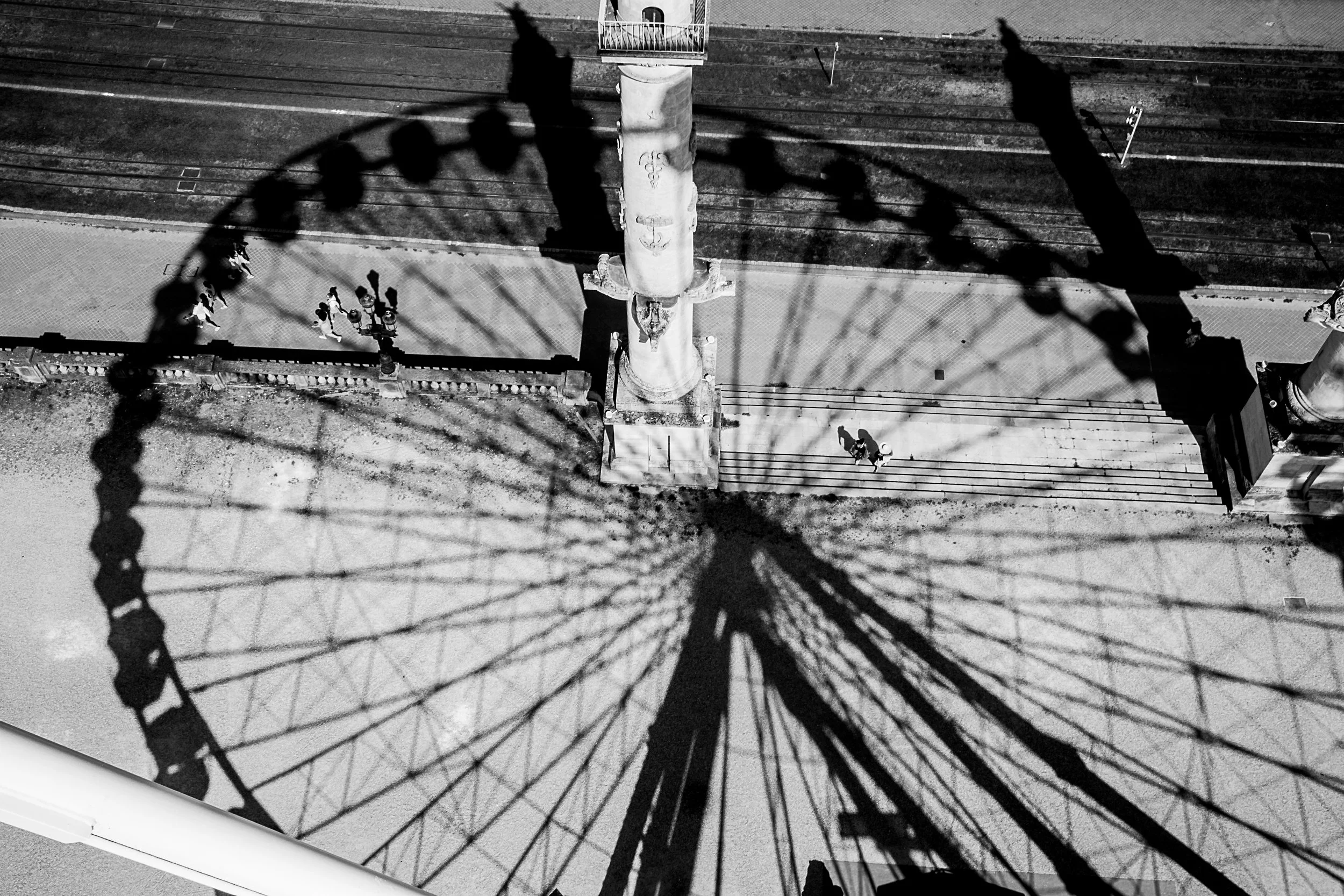 Black and white photo taken from below a Ferris wheel, with children walking near the base of the wheel and a person standing on a balcony above.