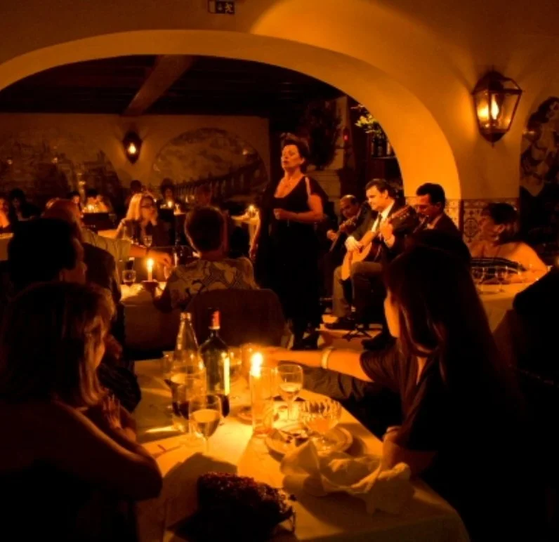 Indoor dining scene with people sitting at tables, candlelit ambiance, live music performance with a singer and guitarists, warm lighting.