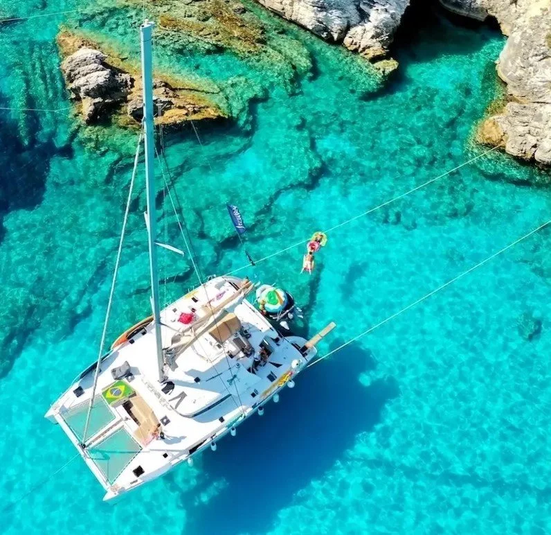 Aerial view of a white sailboat on clear turquoise water near rocky cliffs, with a person swimming nearby.