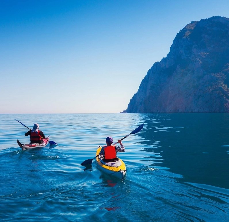 Two people kayaking on a calm sea near a mountainous coastline under a clear blue sky.