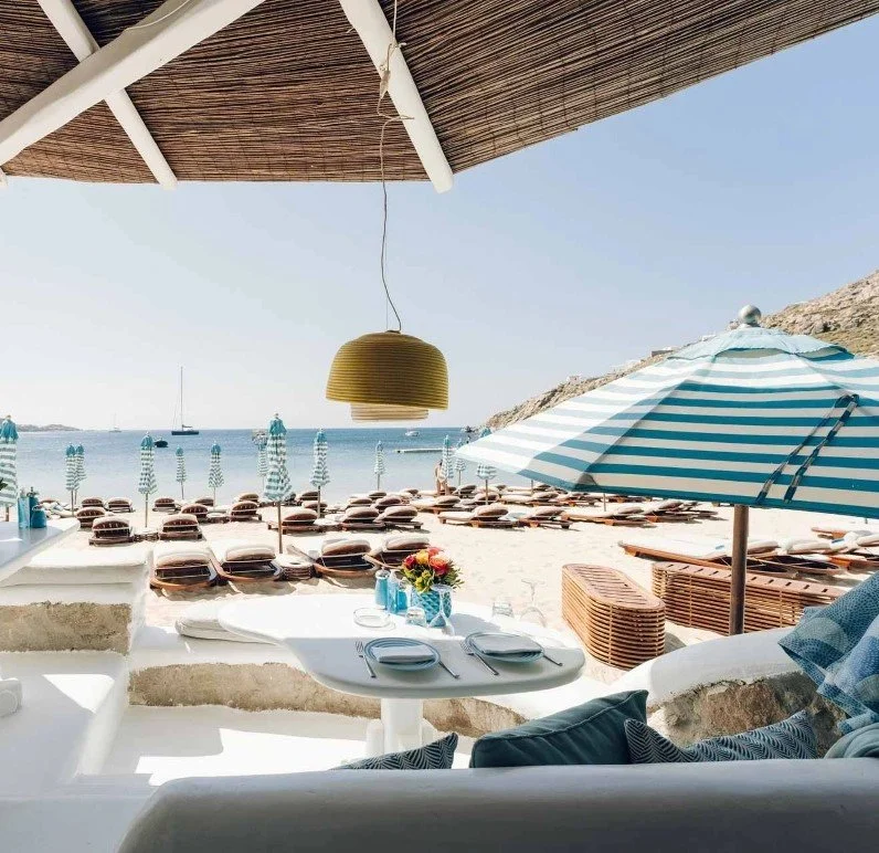 Beachside restaurant with table set for two, overlooking a sandy beach with striped umbrellas and sun loungers. View of the ocean with boats in the distance.